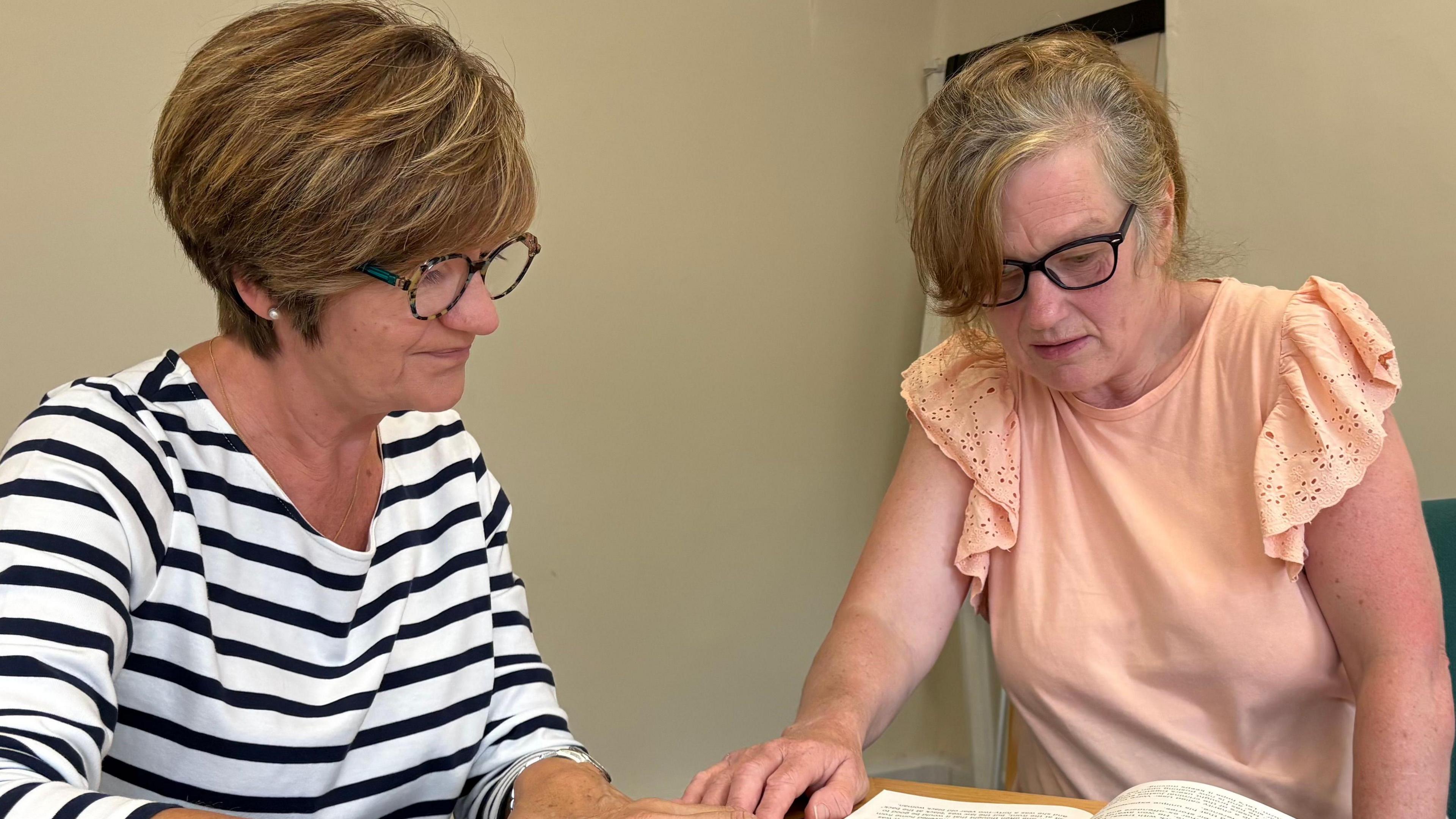 Janet Batson with medium-length brown hair and glasses, wearing a white top with black hoops and Debbie Flaherty, with long brown hair and glasses, wearing an orange top. Both are sitting at a table and looking at a book which has a picture of a woman in it. A paper cup is on the table in the foreground.