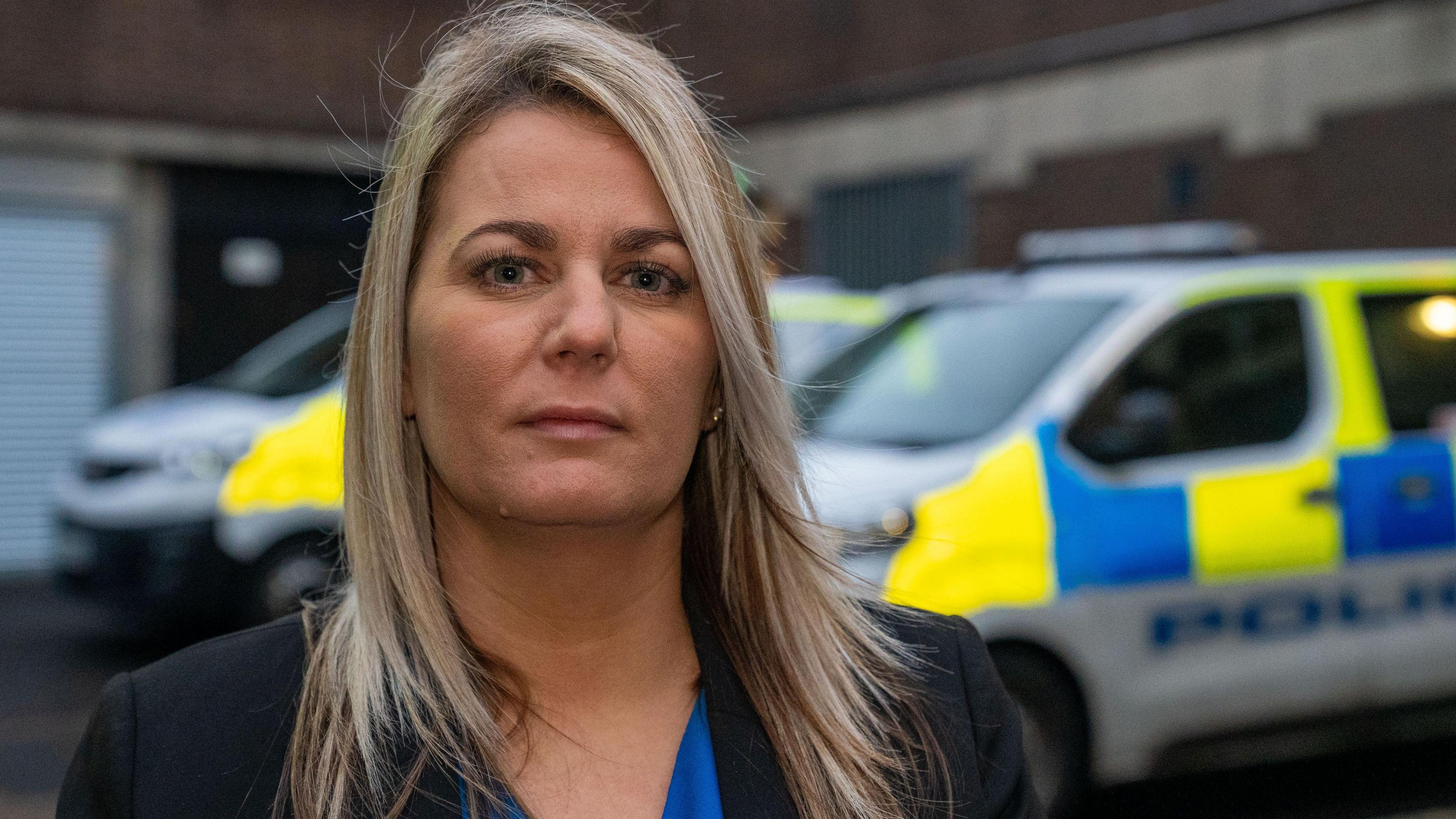 Woman with long blonde hair, head and shoulders shot with police vans in background