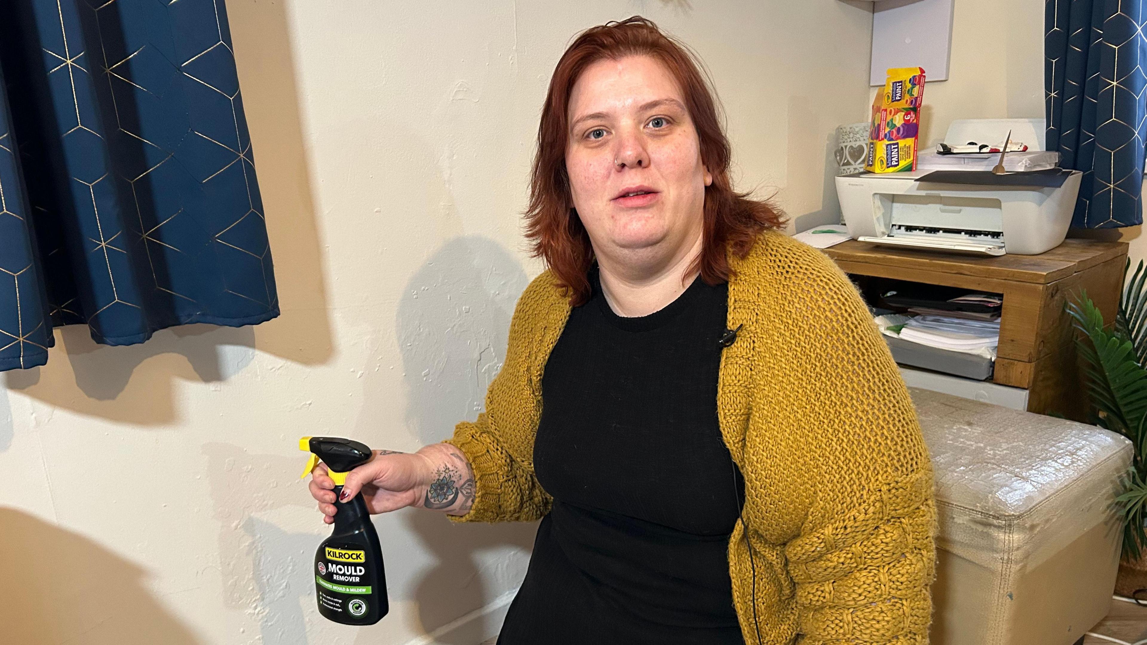 Jade Pack kneels on the floor beside a wall holding a bottle of mould cleaning product. She is wearing a black dress and yellow knitted cardigan. Behind her is a printer on a desk, and to her right are blue curtains. 