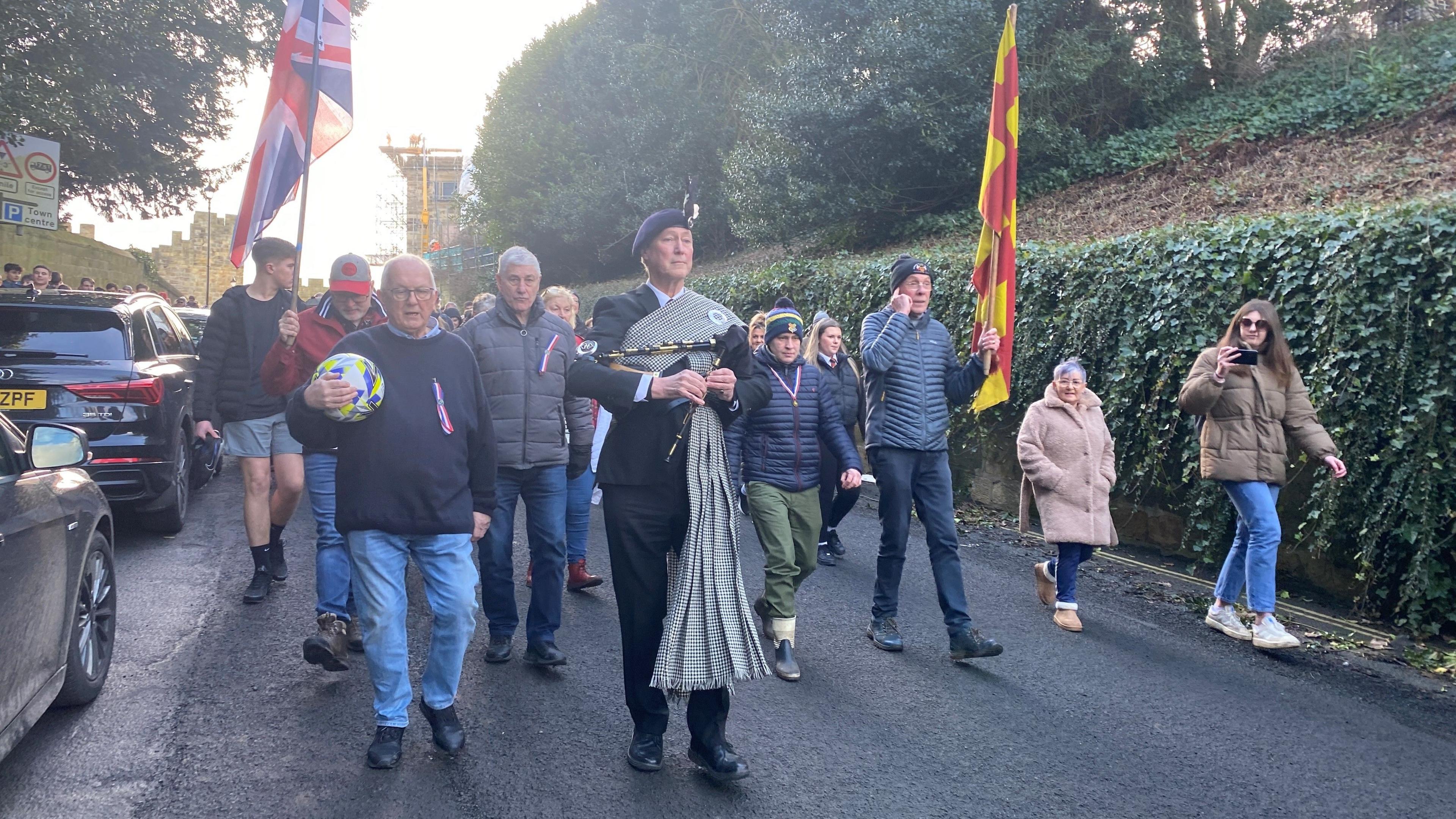 Dozens of people are pictured walking down a road, led by a piper. One man is carrying a ball, another - to the right - is carrying the red and yellow flag of Northumberland, and a third man - to the left - carries the Union Jack.