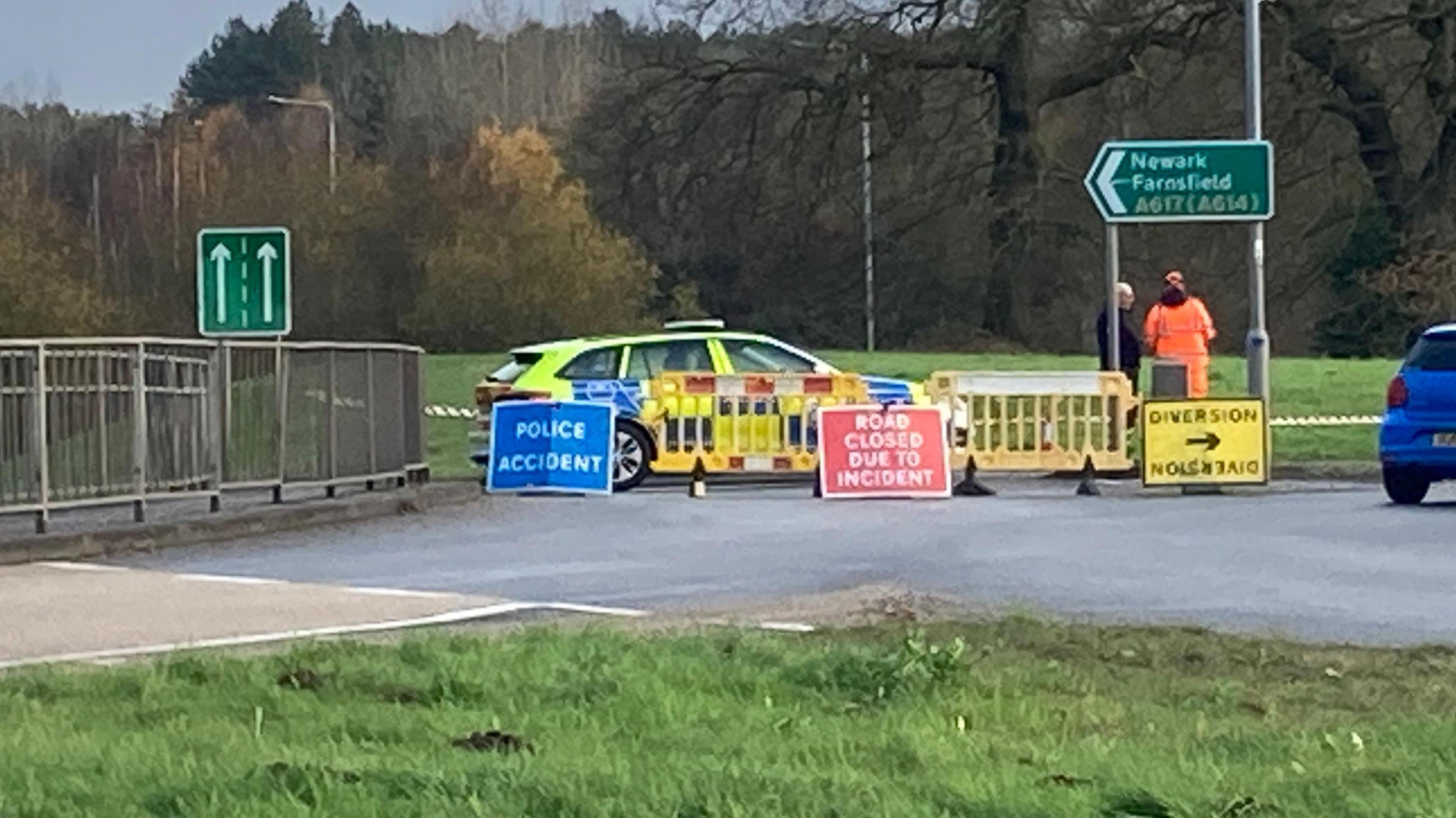 A police car at the A617 in Rainworth, Nottinghamshire. The car has green and blue markings and is behind temporary yellow plastic fencing. Three signs stand in front. A blue one on the left has "police accident" written on it in white letters. The middle one, a red one, has "road closed due to incident" also in white letters on it. The right hand one is yellow with black wording saying "Diversion" with an arrow pointing to the right. Two people stand to the right of the police car next to a sign for Newark.