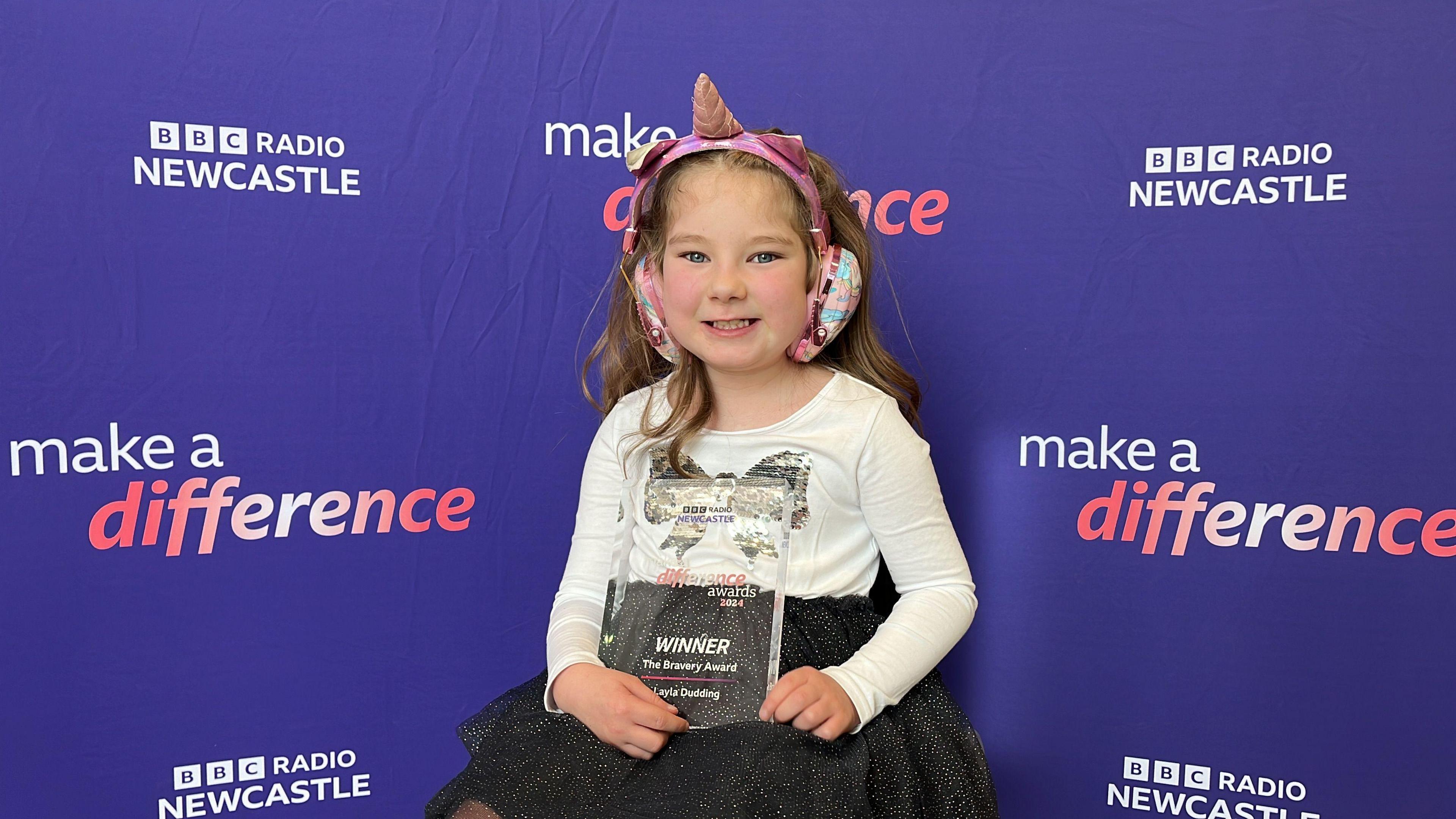 Layla Dudding. She is seated and holding her award. She is wearing a white top, black skirt and is wearing ear defenders with a unicorn horn attached.