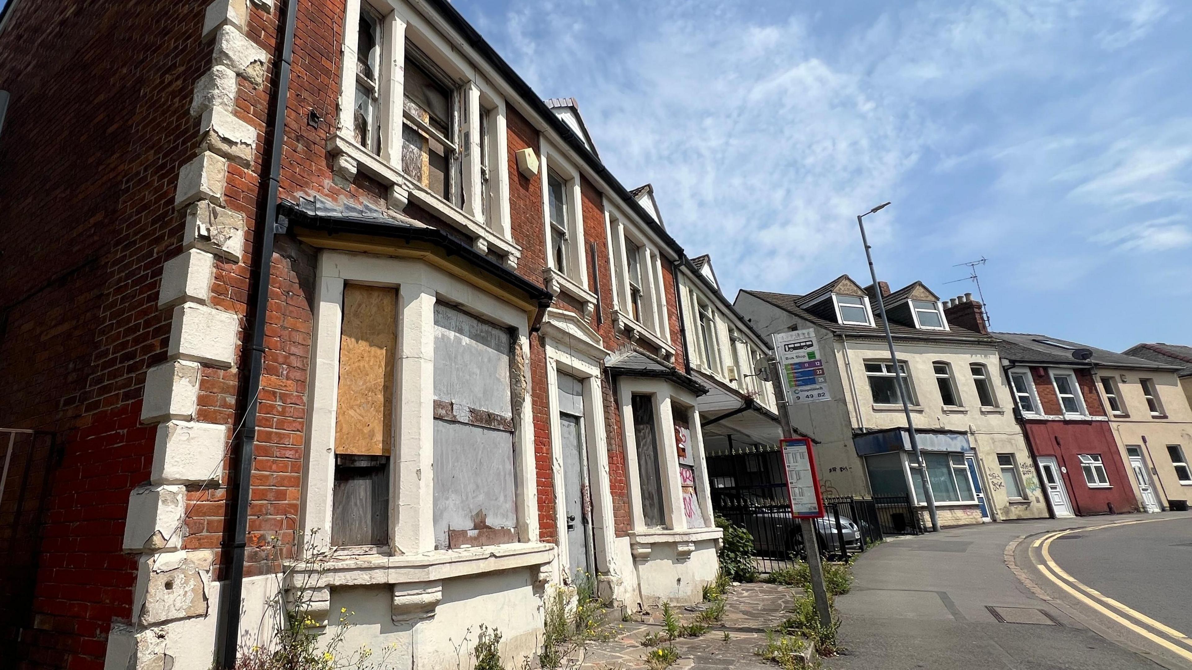 An empty home which has its windows boarded up because they are smashed. It's door handle is hanging of. Paint is peeling away. Weeds are growing out of the floor. It sits on a residential road.
