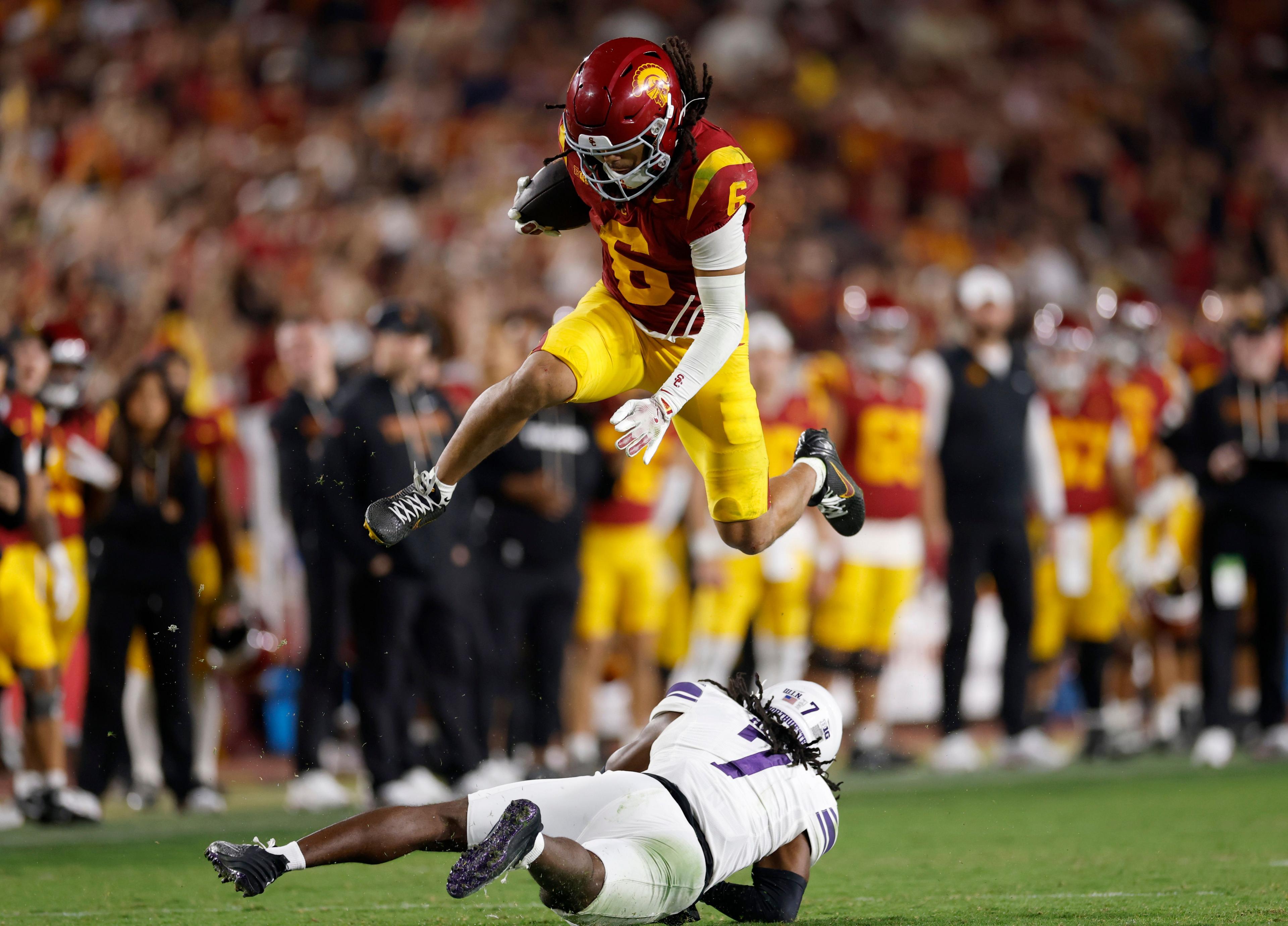 A football player in a red and yellow uniform leaps over an opposing player in a white and purple uniform who is lying on the ground during a game, with a crowd of spectators and teammates visible in the background.