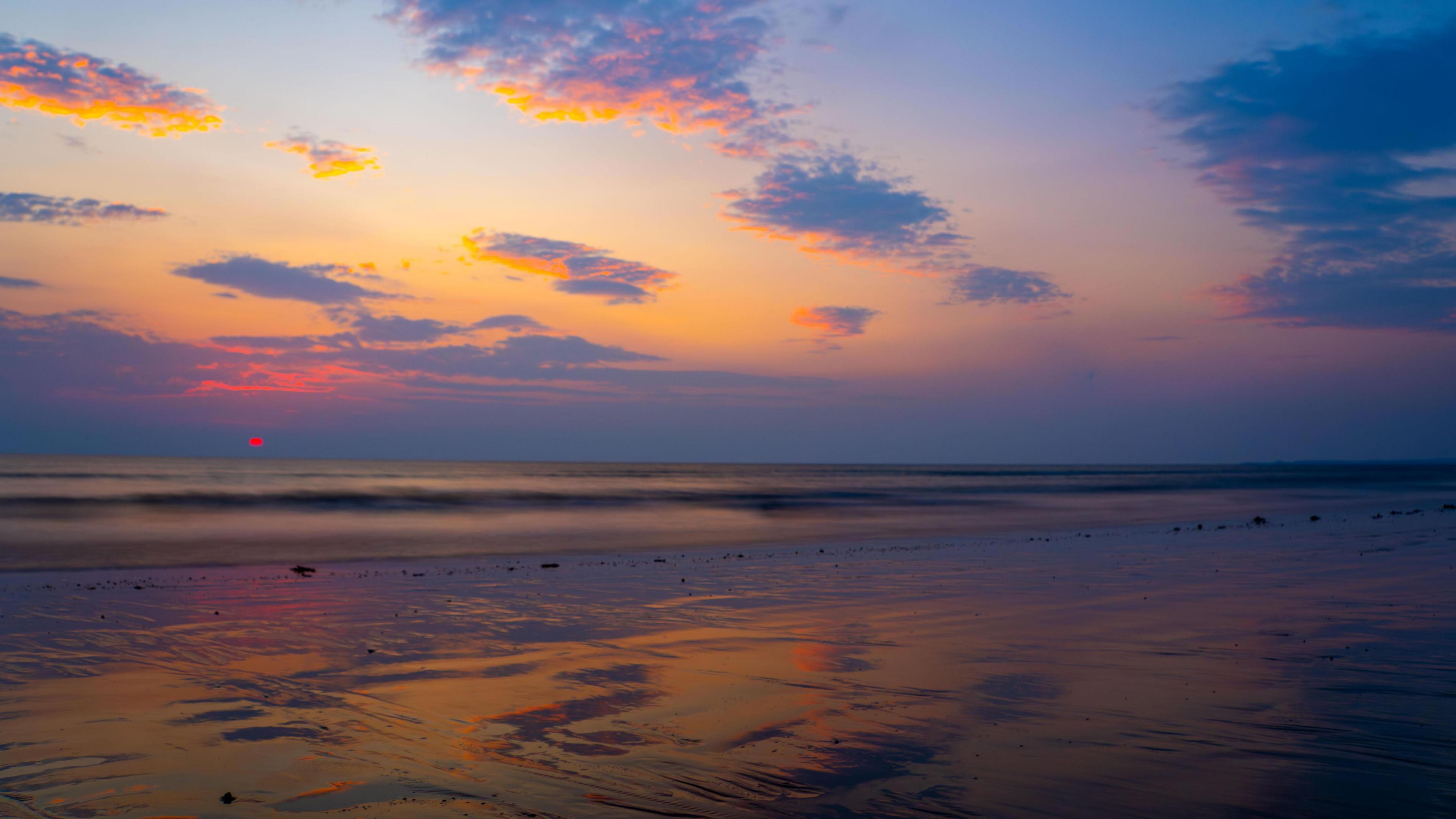 Sunset on a beach looking out to sea with some dark orange colours on the horizon, and a bit of cloud overhead with a tinge of yellow and orange