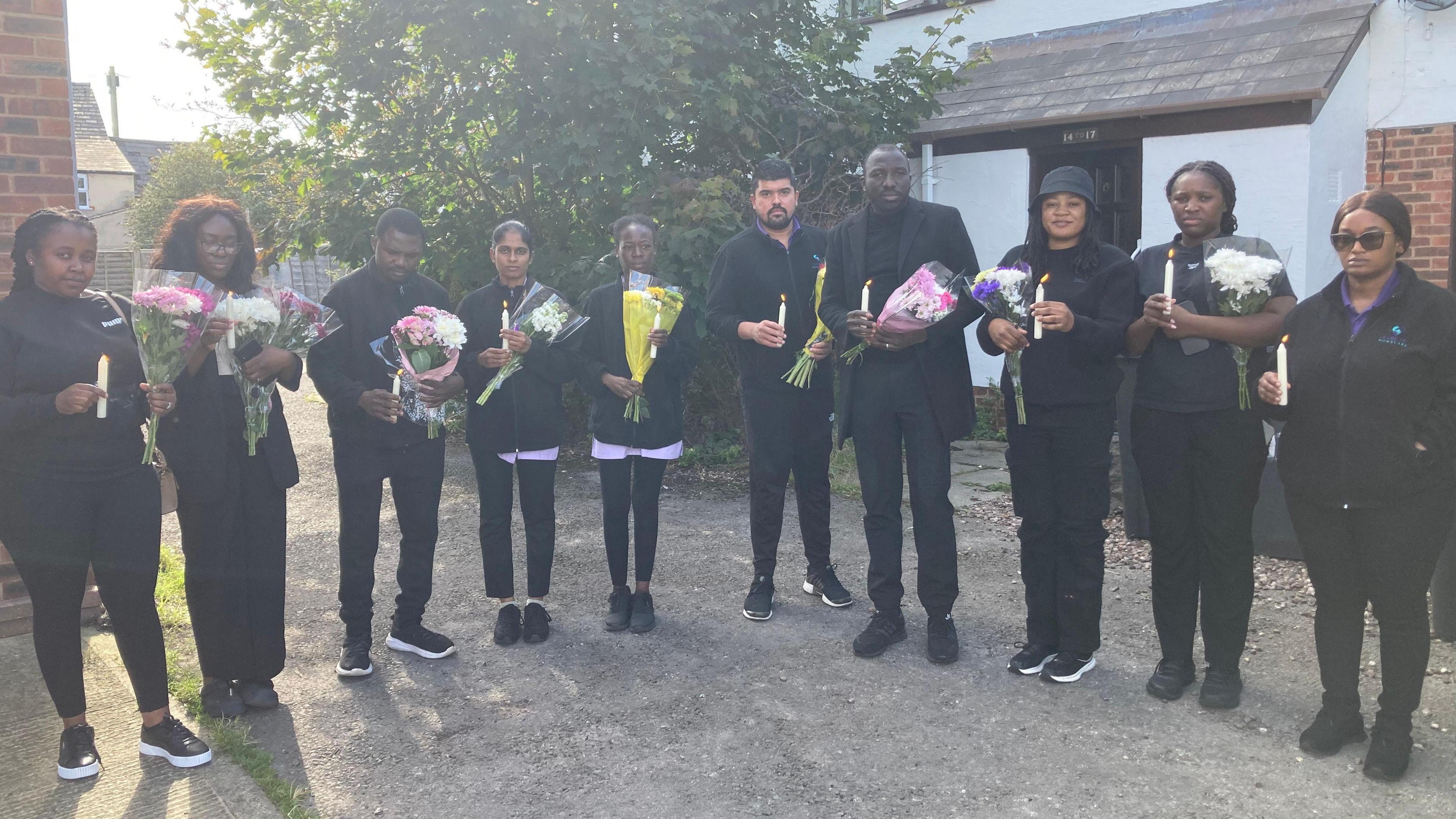 10 people holding lit candles and flowers at a vigil outside a house