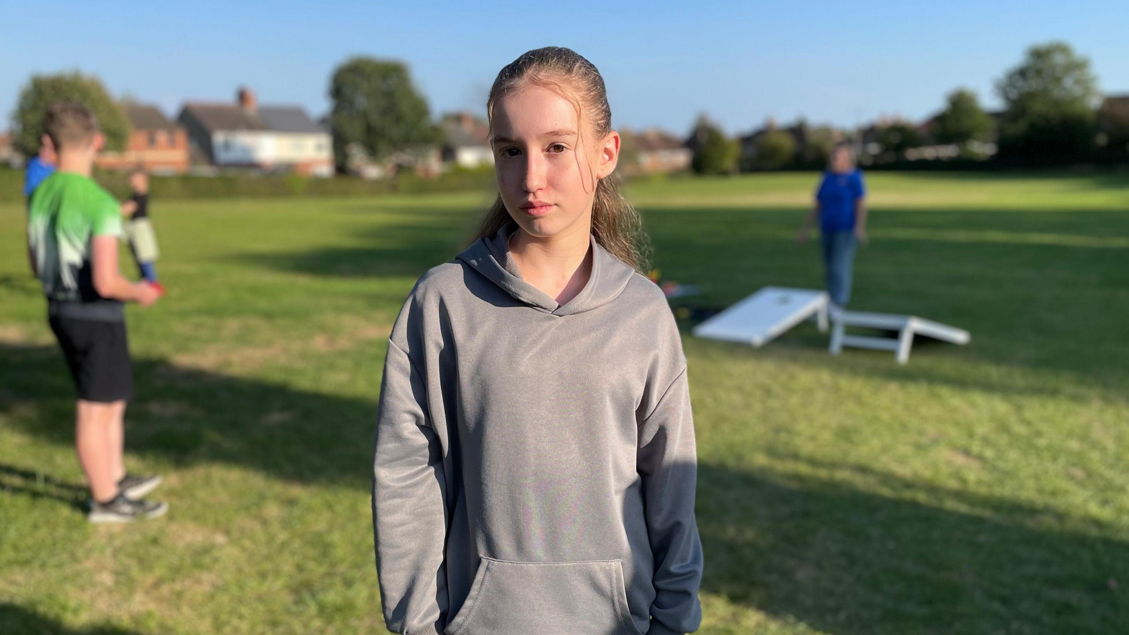 Ella, 13, is pictured in a grey hoodie with long dark hair which is tied back. In the background, youths who are blurred play a game.