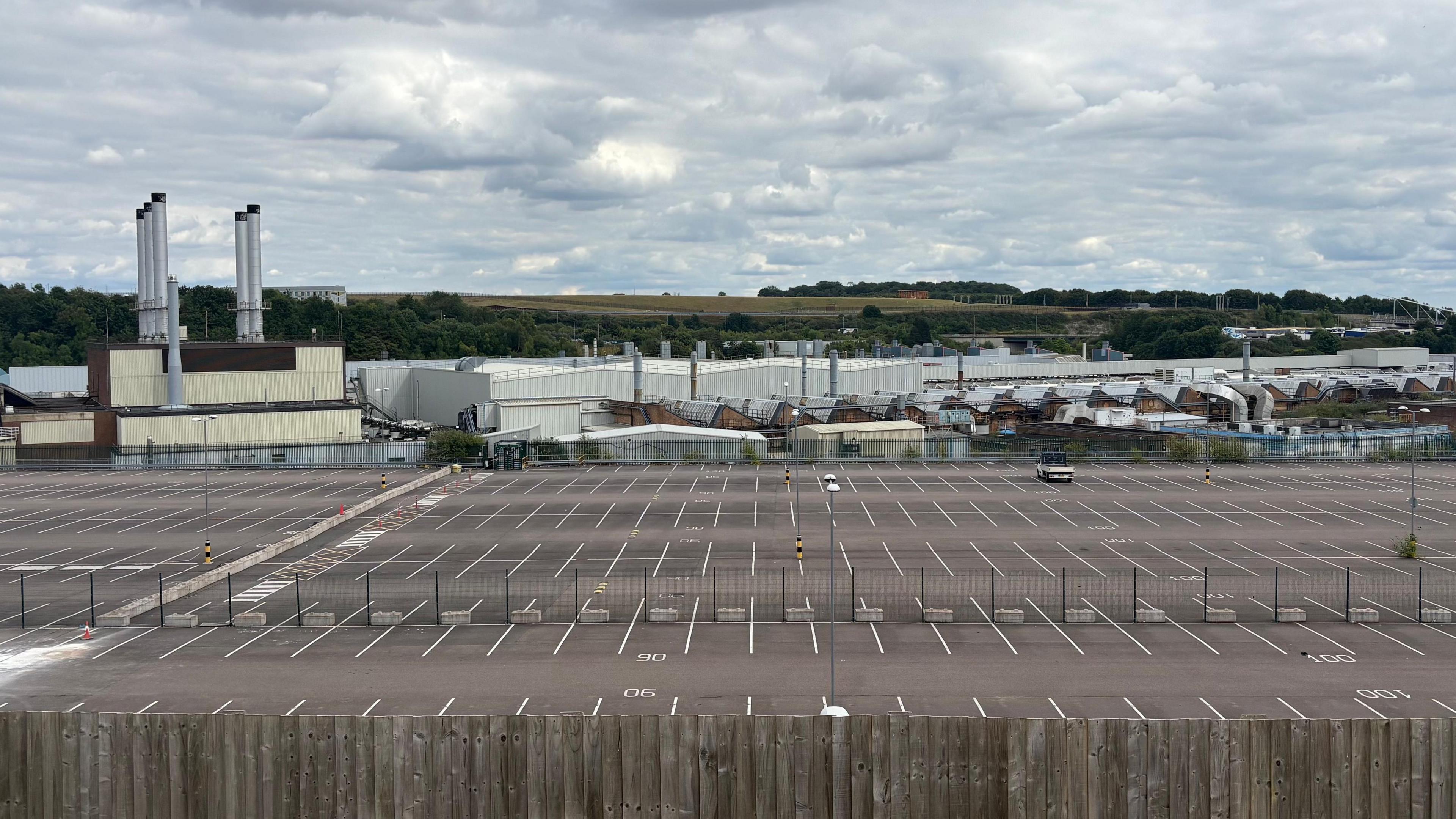 A wide shot of the Vauxhall factory in Luton. In the foreground is a large surface, open air car park, which is empty, except for one truck in the middle distance. Beyond the car park is a set of low-rise industrial buildings. On the left, five metallic chimneys stretch into the sky. Behind the buildings is a hill covered in grass and trees.