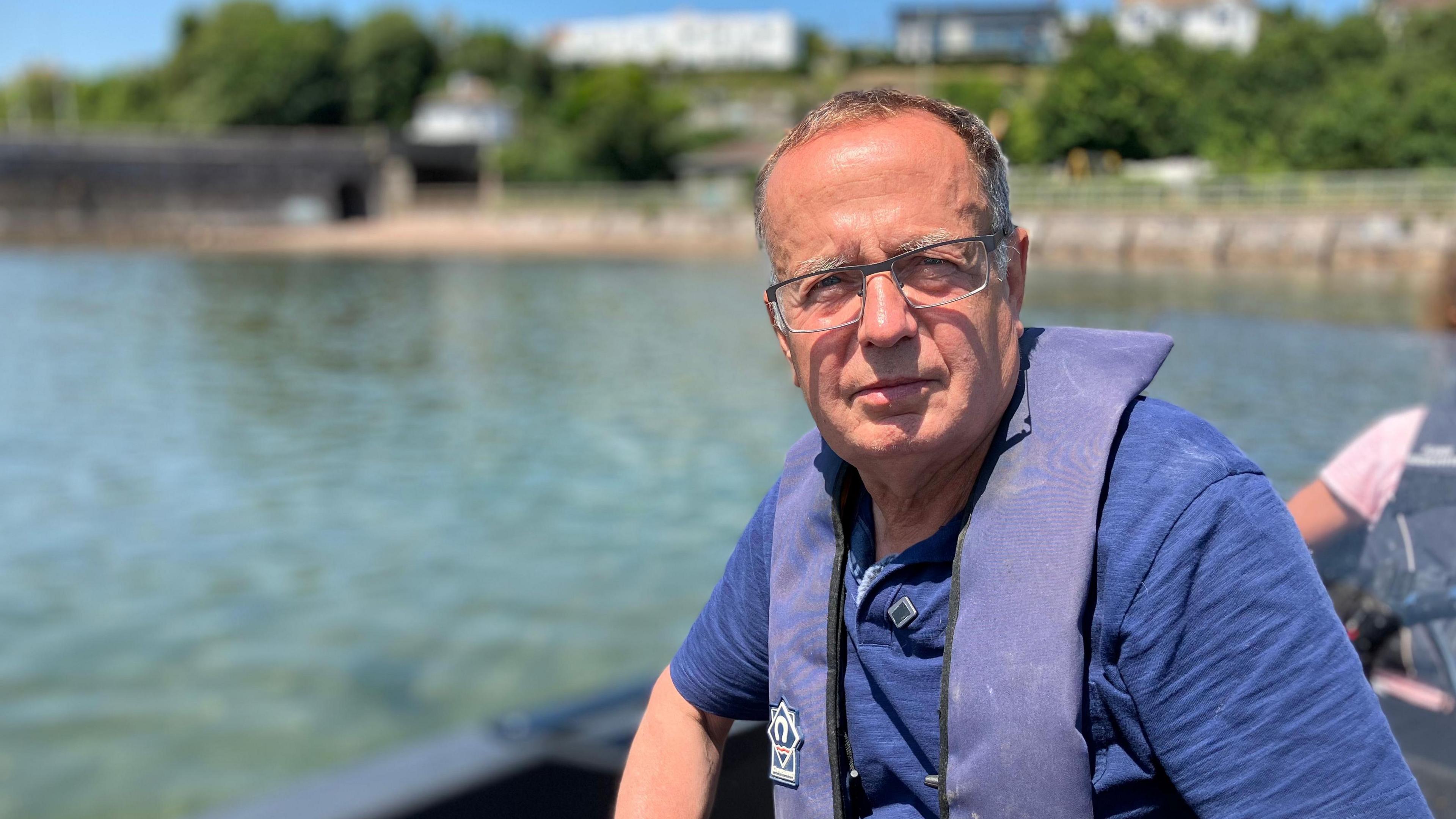 A man sits in a small boat in a river estuary. He is wearing a blue t shirt and a blue life jacket. The sun is shining and reflecting off the water.