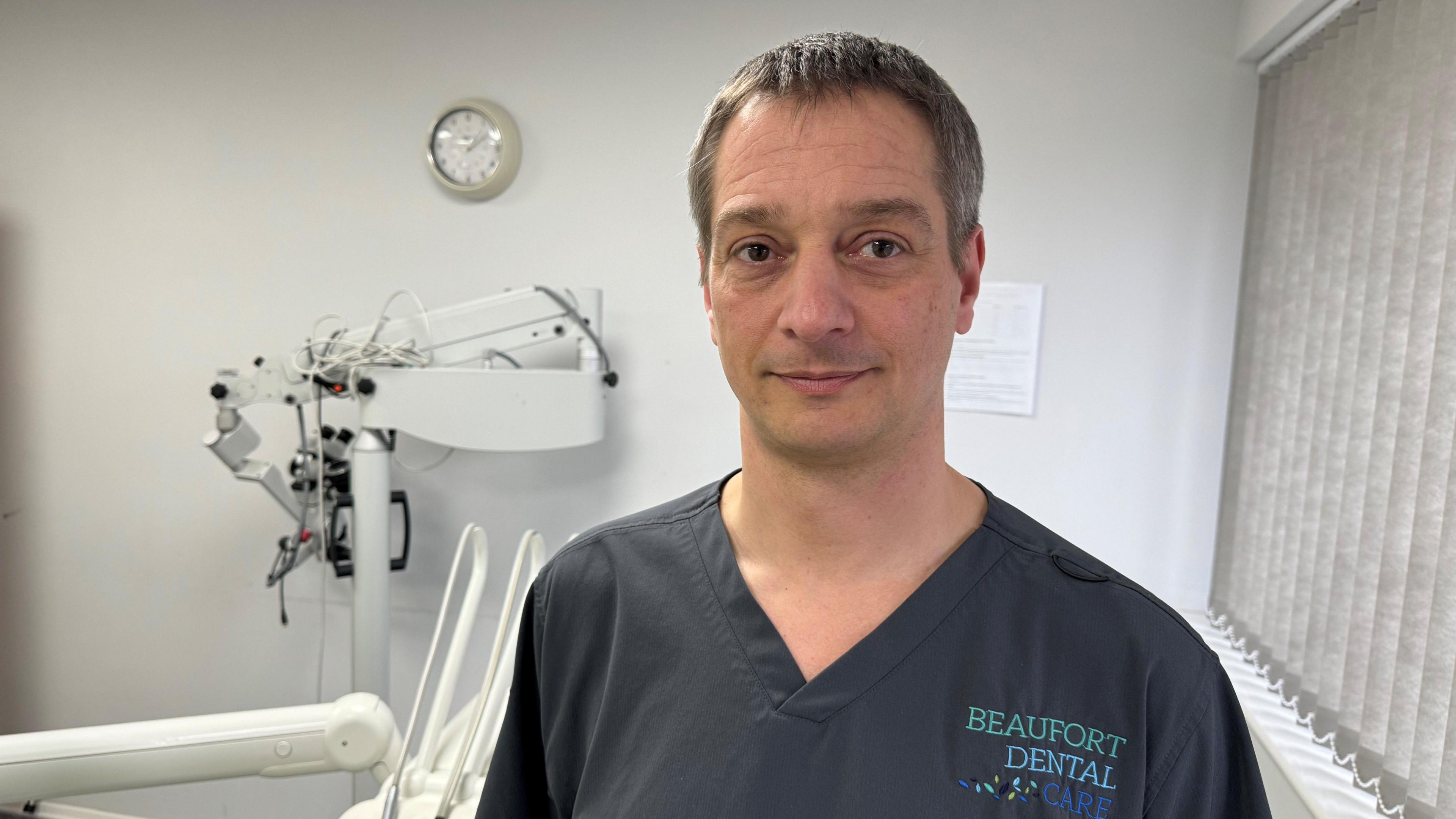 Russell Gidney is wearing his dental scrubs. he is posing for the picture in his dental practice clinical room.