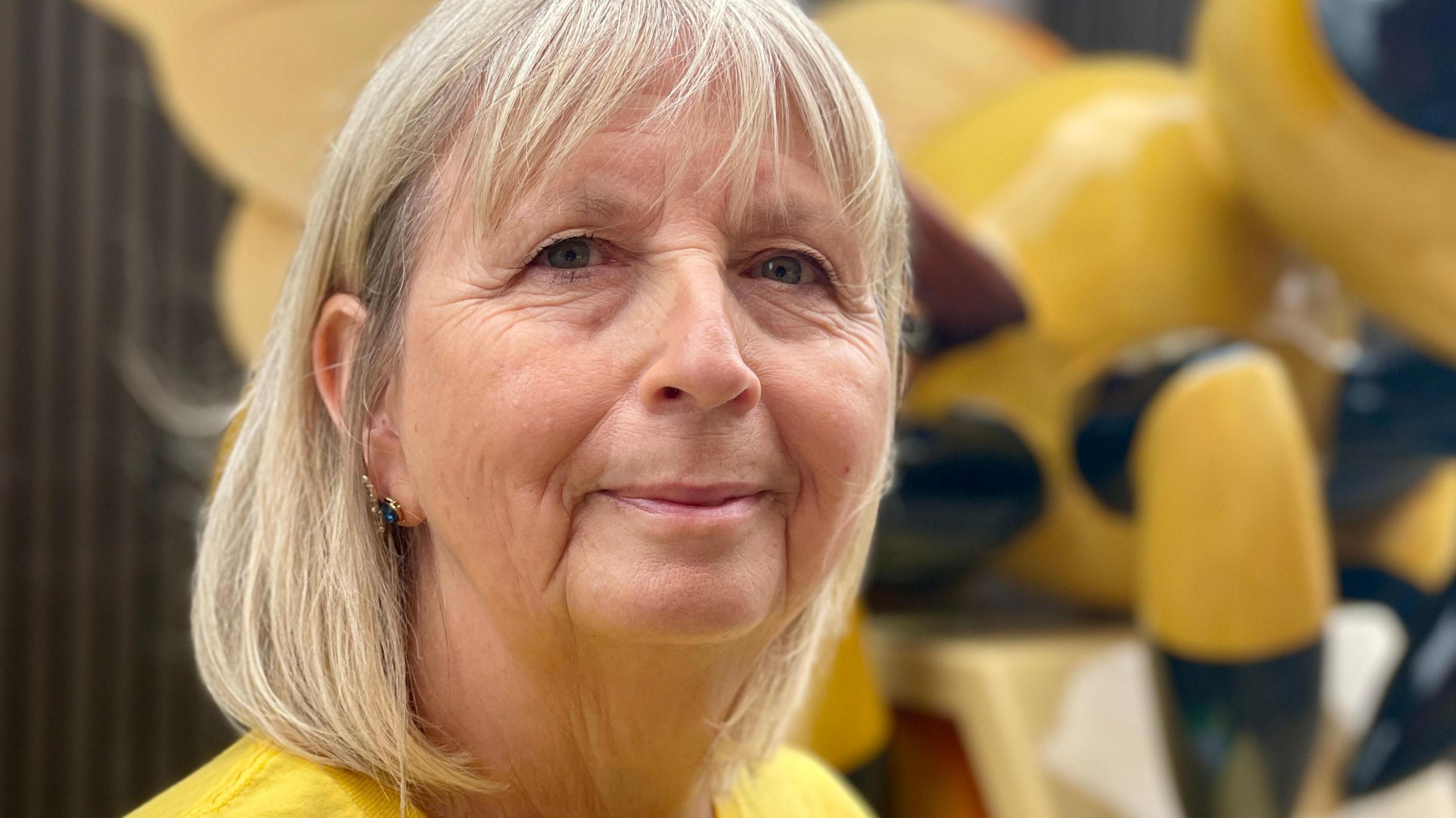 Pauline Harvey-Jones has a blonde bob, and is wearing a yellow T-shirt. Part of a yellow and black bee sculpture can be seen behind her.