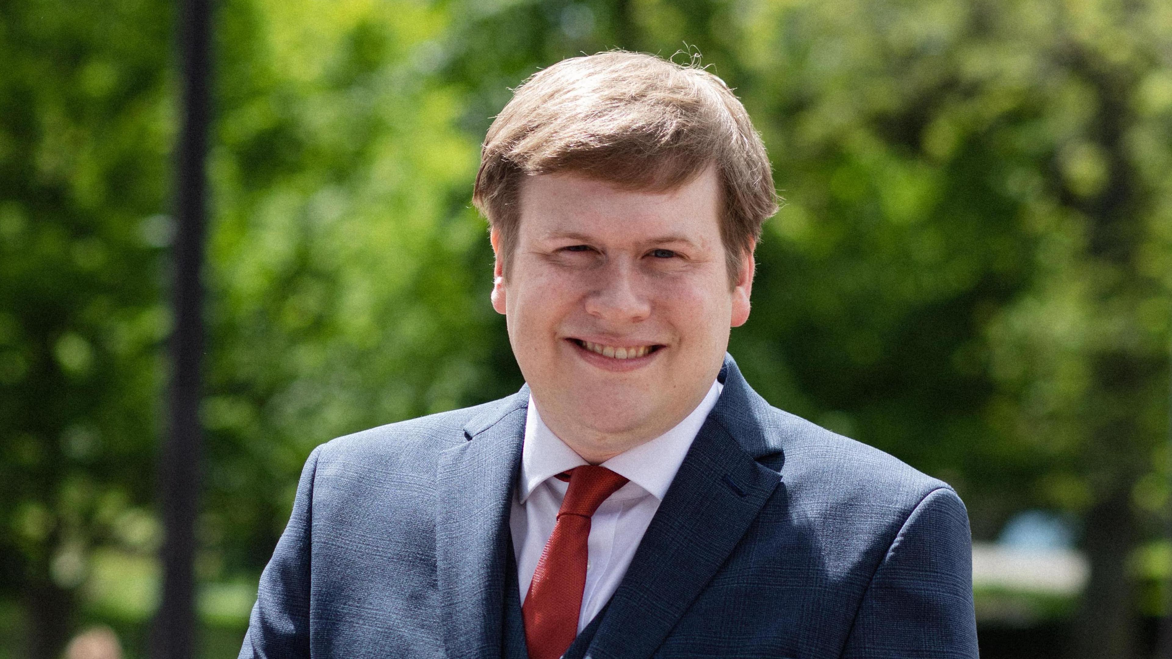 Alex Hay has light brown hair with a side parting. He is wearing a blue suit with slight check pattern and a red tie. He is smiling directly at the camera.