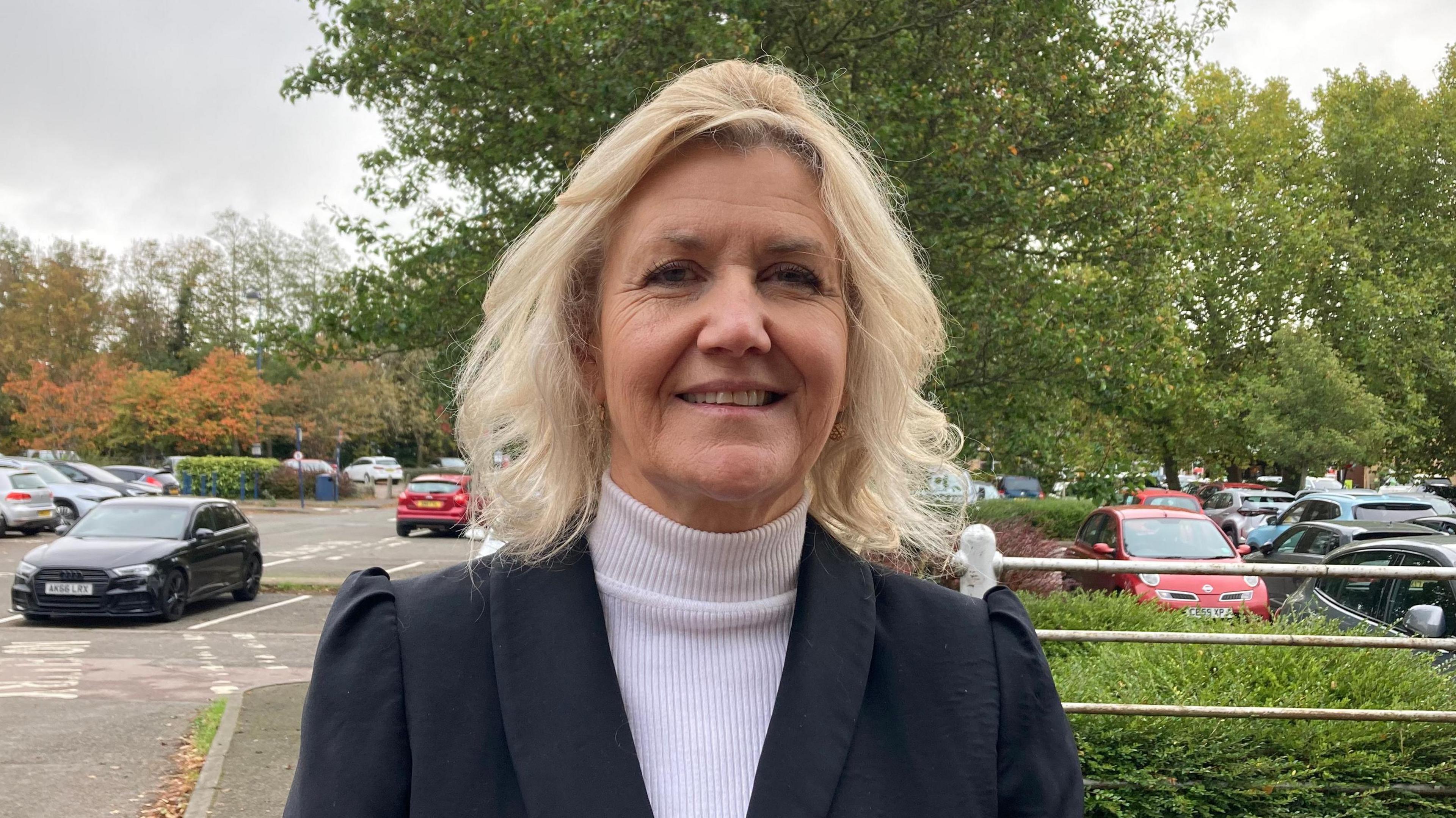 Close up image of Julie Kerr. She wears a dark jacket with a white top. She has shoulder length blonde hair. Behind her is a car park with a number of trees and bushes