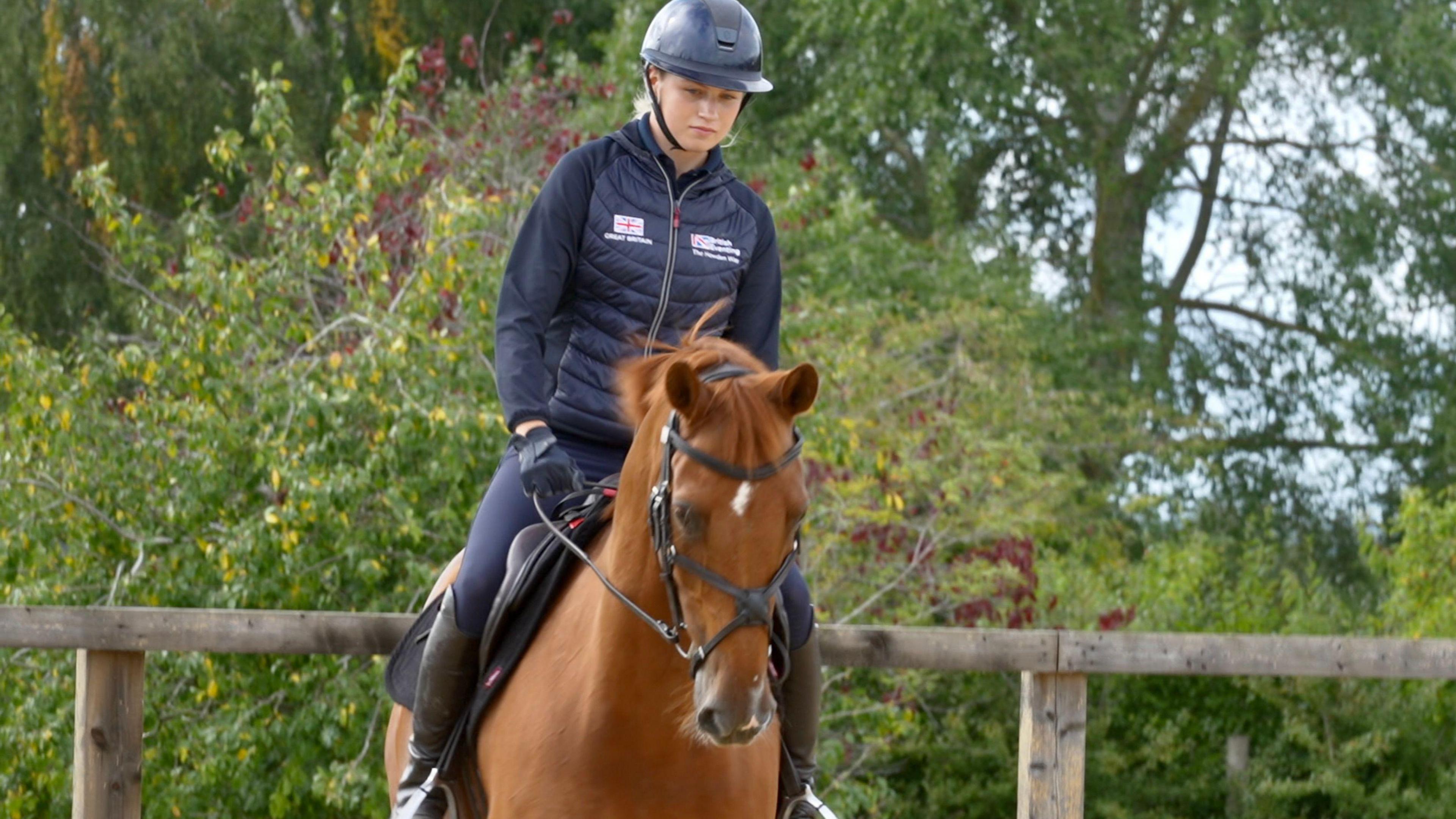 Ella rides Spice the horse in a training outdoor school. She wears a navy helmet, puffer coat, globes and black riding boots.