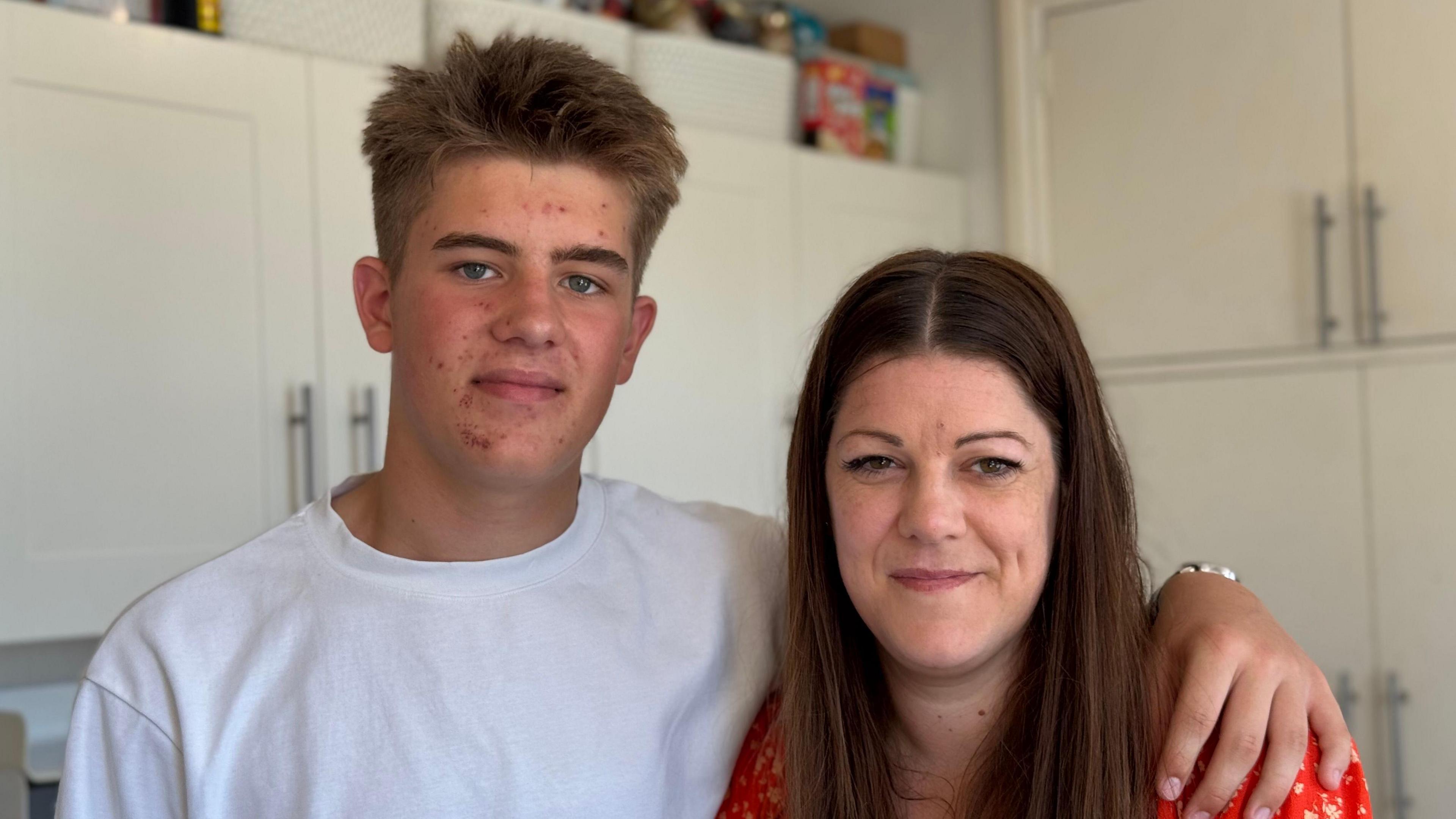 Oliver and his mum Wendy hug one another. On the left, Oliver has short spiky hair and wears a white t-shirt. On the right, Wendy has ling brown hair and wears a red dress with a white pattern.