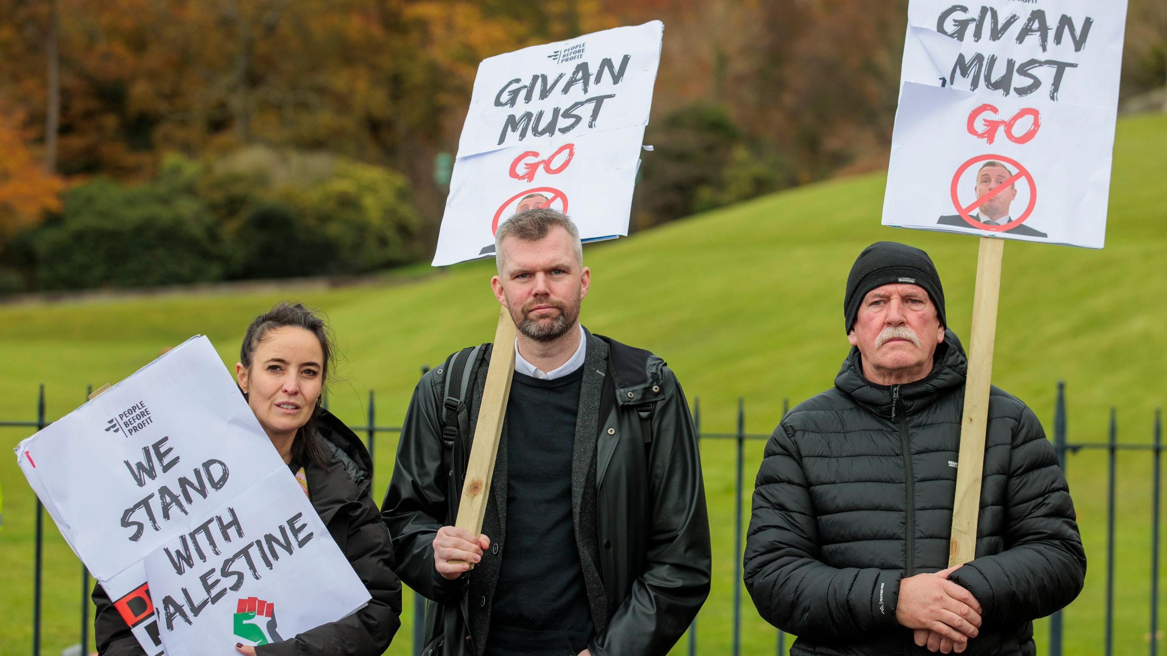 Gerry Carroll stands with two protesters, a man and a woman, during a demonstration at Stormont. They are all wearing dark-coloured coats and carrying placards. Carroll, who has short, greying hair and beard, is carrying a "Givan Must Go" placard, as is the man beside him who has a grey moustache and a black beanie hat. The woman, who has dark hair tied back, is holding a "We Stand With Palestine" placard. There are trees and a green lawn in the background.