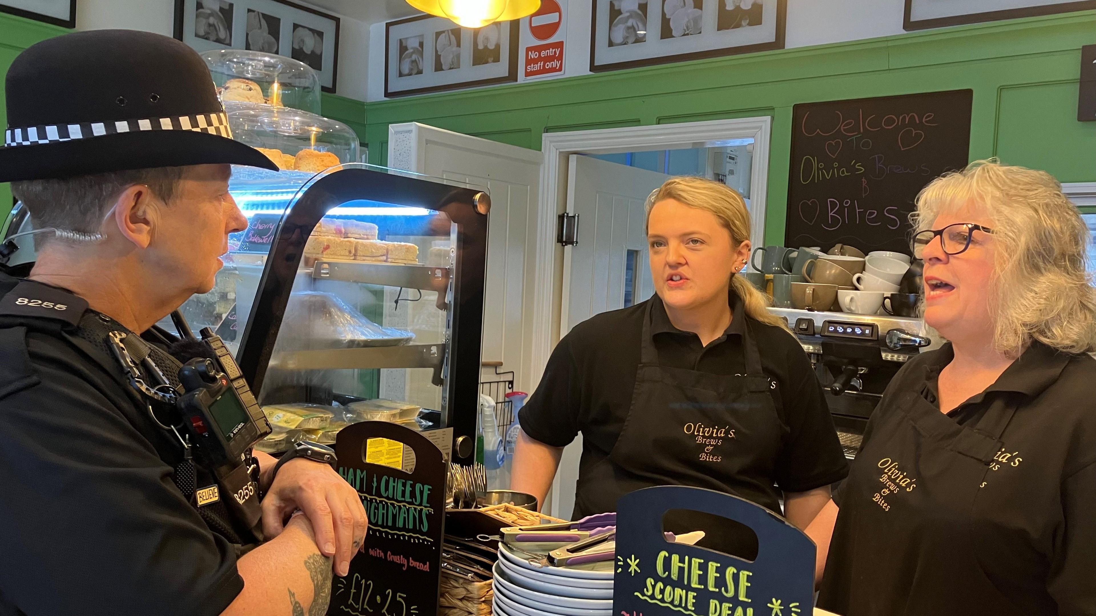 PC Pip Davey is dressed in full uniform standing in front of a shop counter talking with Olivia and Jo Richards who are dressed in a shop uniform. There are coffee cups stacked behind them and a counter display with sandwiches and cakes and small blackboards detailing special offers including a cheese scone deal.