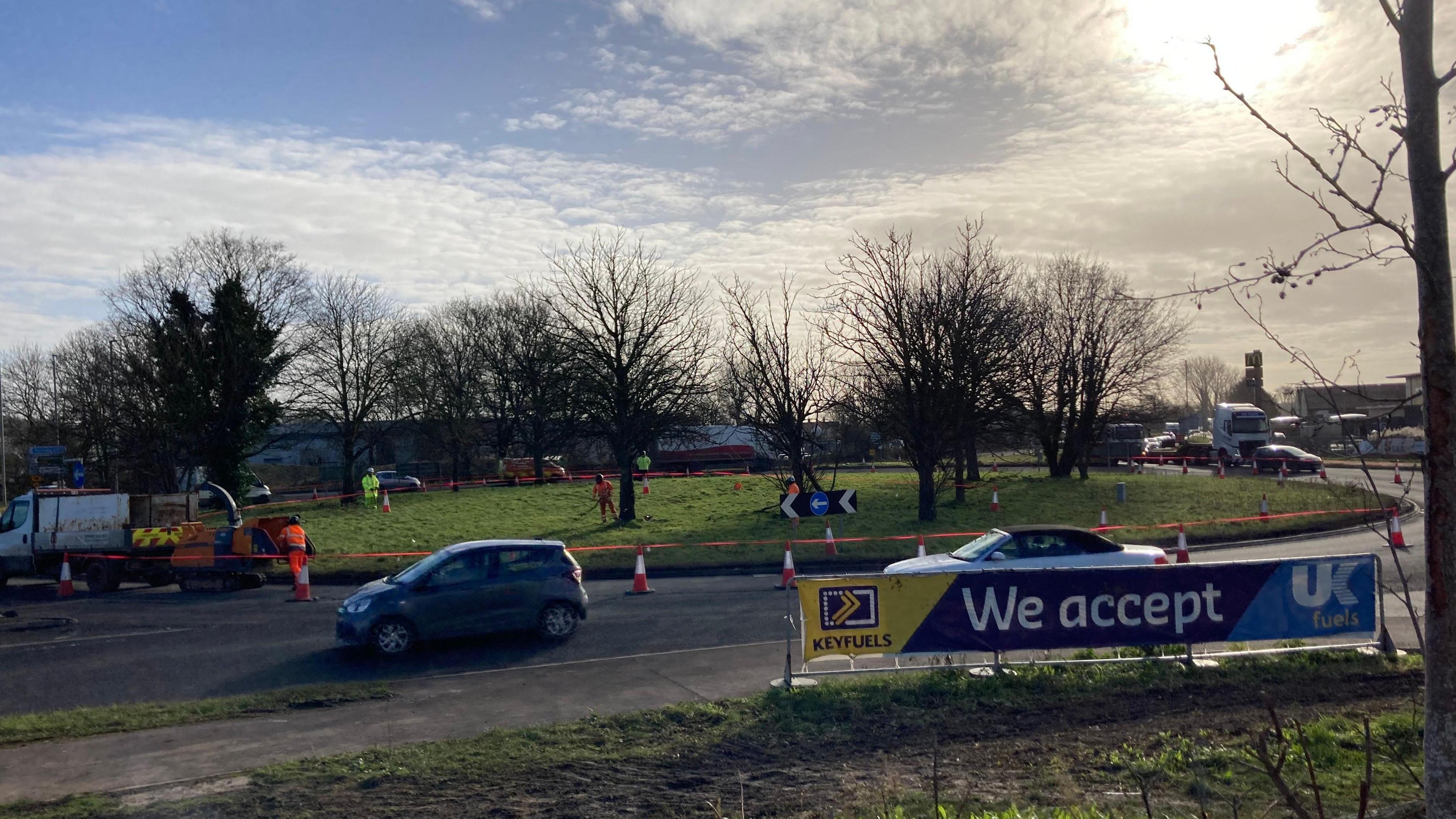 Roundabout with green grass in the middle with cars going around it. There is cones and a orange tape with roadworks ongoing.