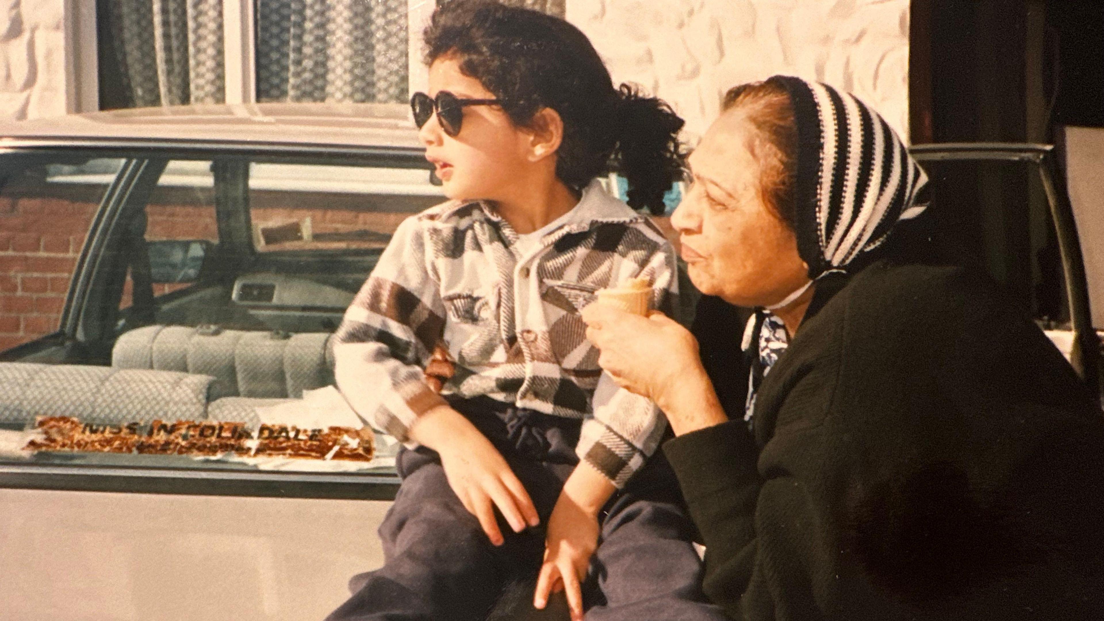 Old photo showing Pavan as a child, with long dark hair and wearing sunglasses, sitting on a car boot next to his grandmother, who has dark brown hair and is wearing a headscarf