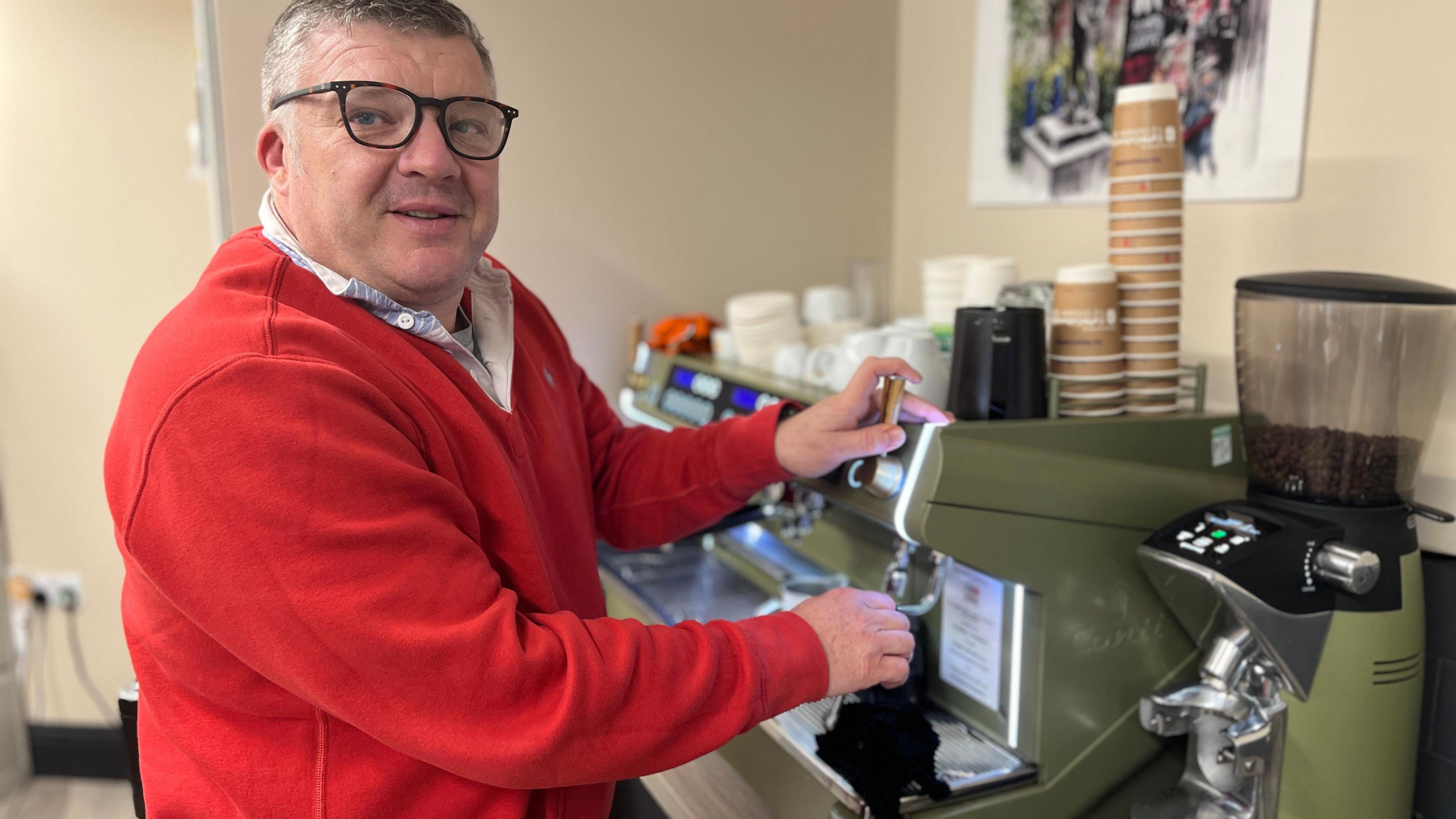 Nigel Seaman, wearing a red jumper, stands using a coffee machine inside an office.