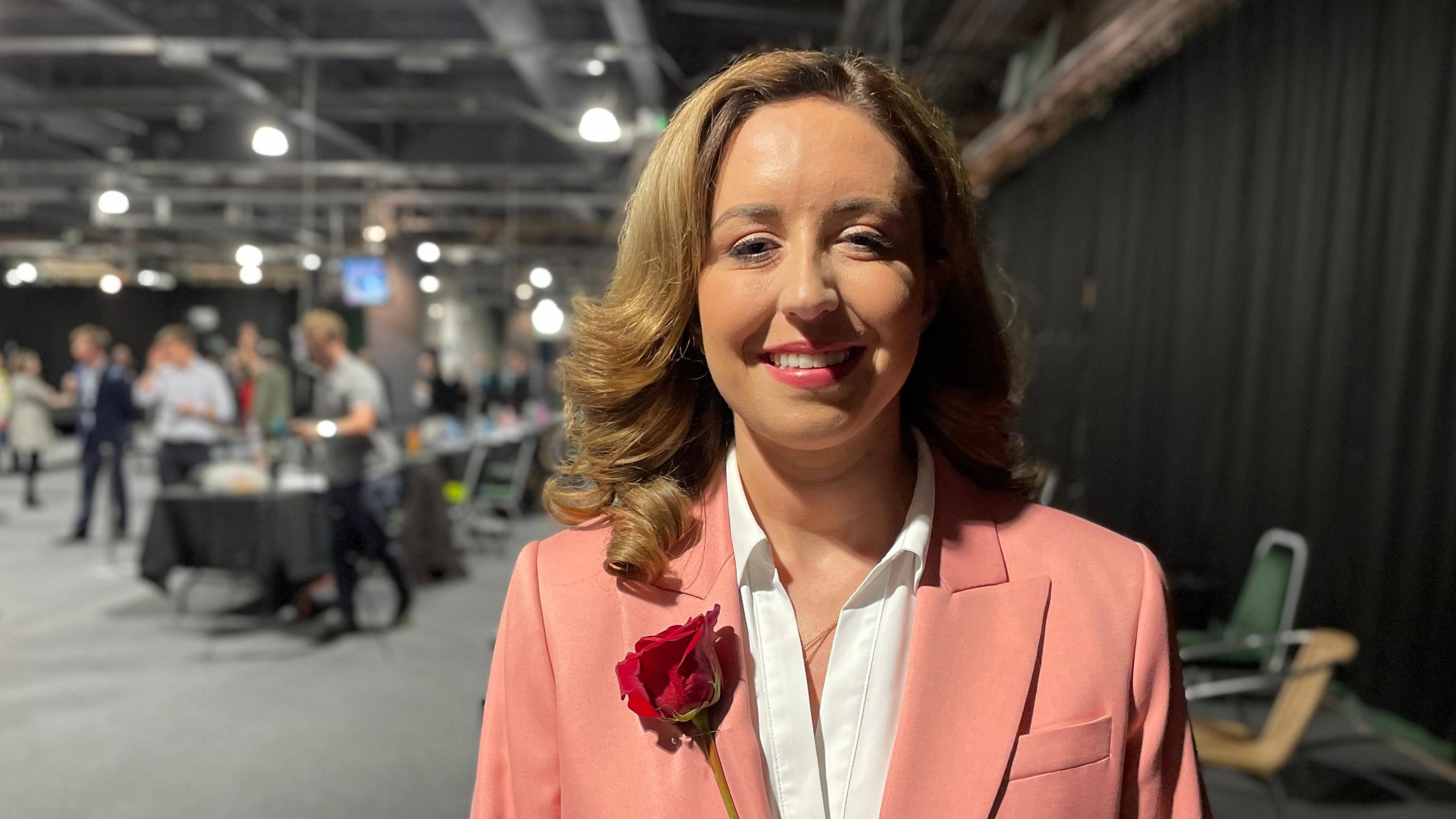 Sally Jameson stands in a hall. She wears a salmon-coloured blazer and white blouse with a rose fixed to her right side. She has curly, shoulder-length brown hair.