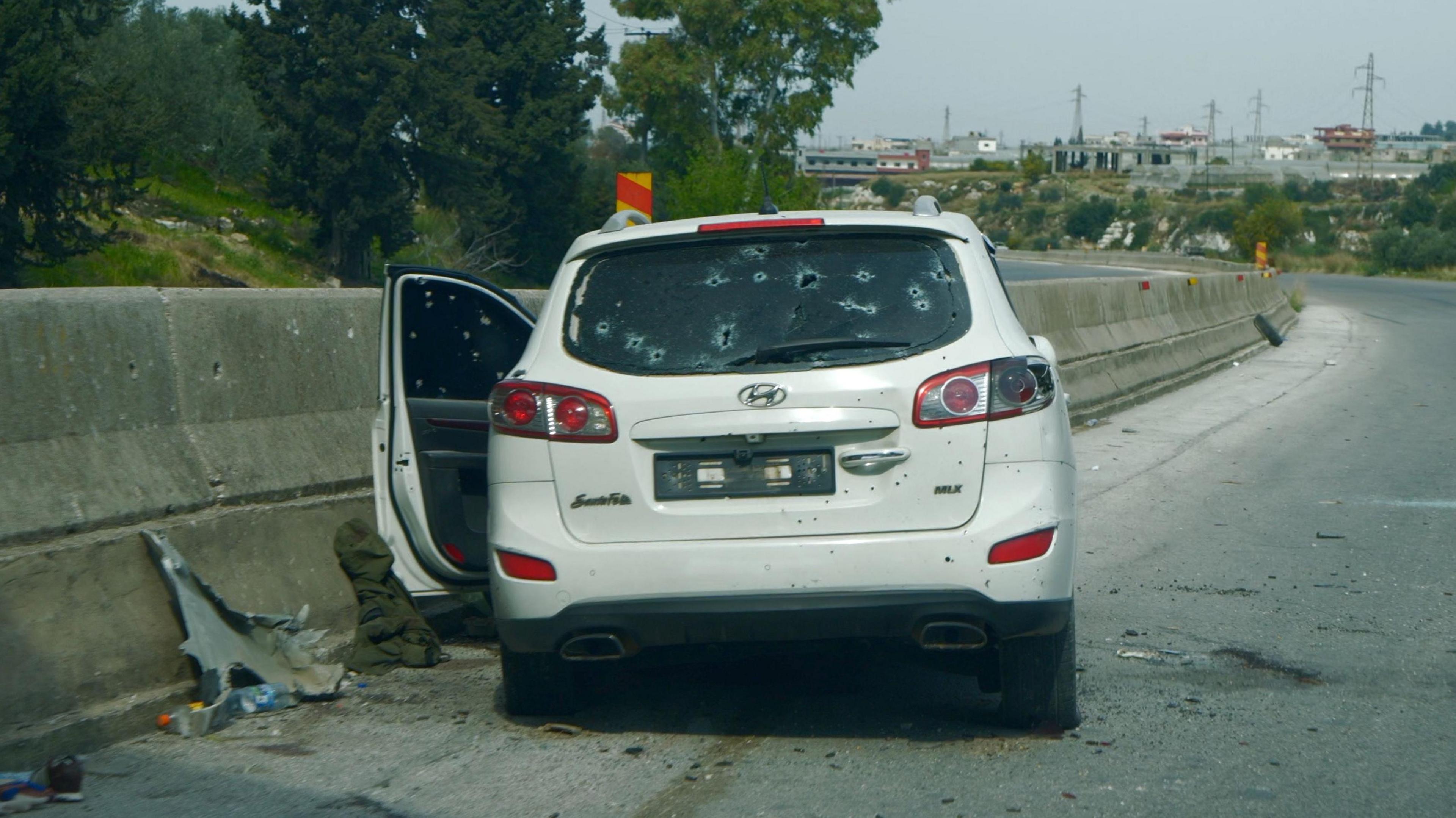 A white car with bullet holes sits against the central barrier on a highway. One door is open, and there is clothing on the ground. There are no people visible in the picture 