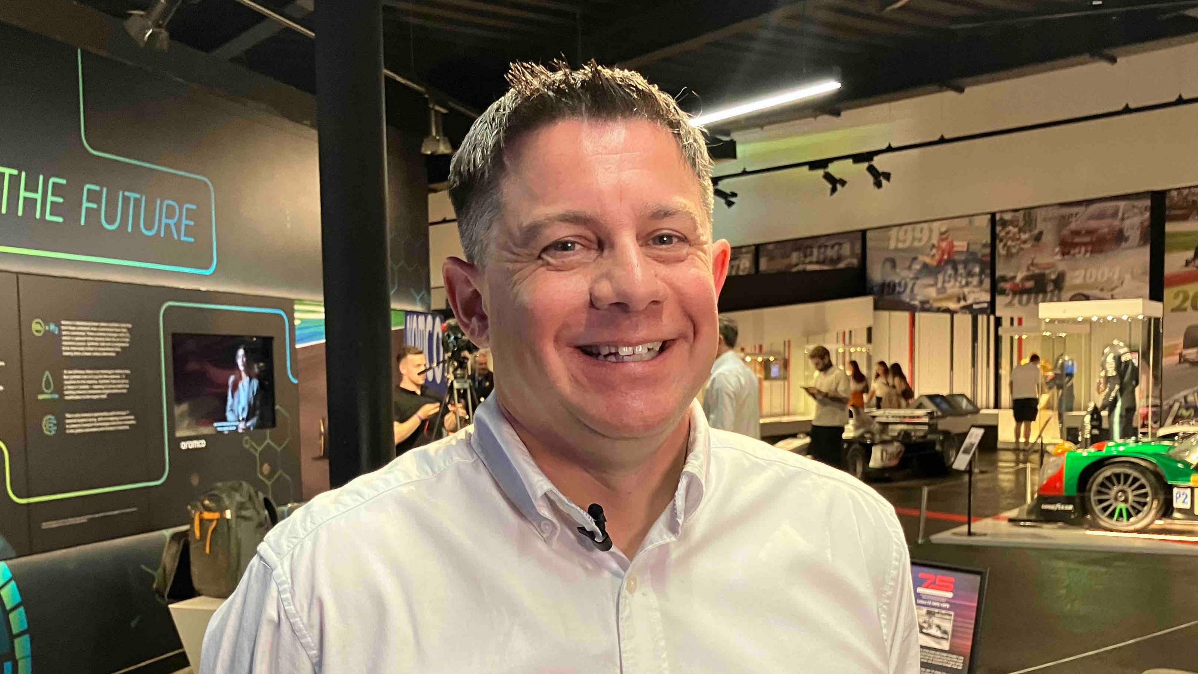 Rob Jaina with short blond hair smiling at the camera and wearing a white shirt. There is a display behind him called "The Future" and part of a green car is visible to his left. There are people looking at exhibits behind him.