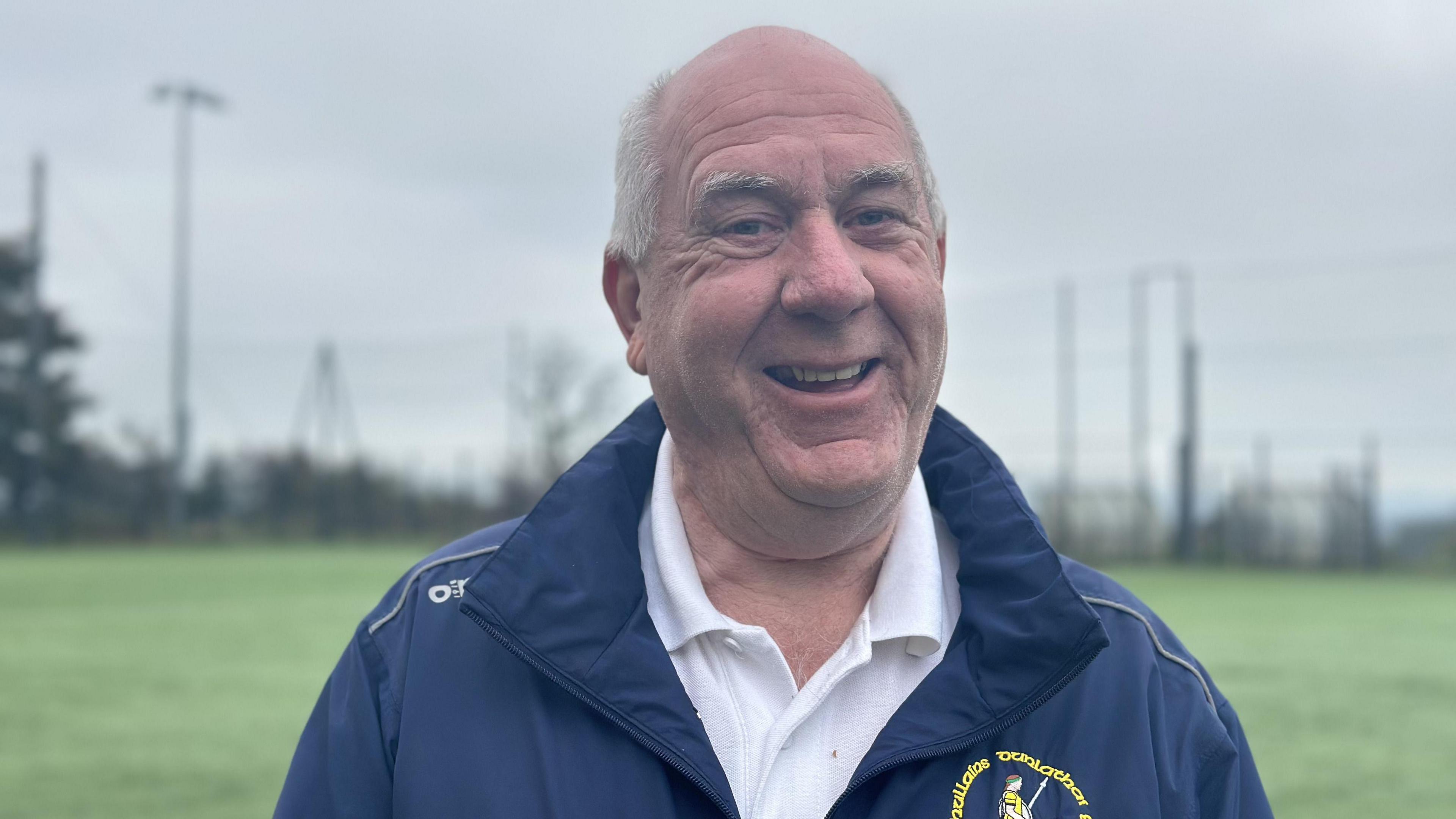 An older man wearing a white shirt and navy GAA coat. He is smiling at the camera and is standing in front of a GAA pitch.