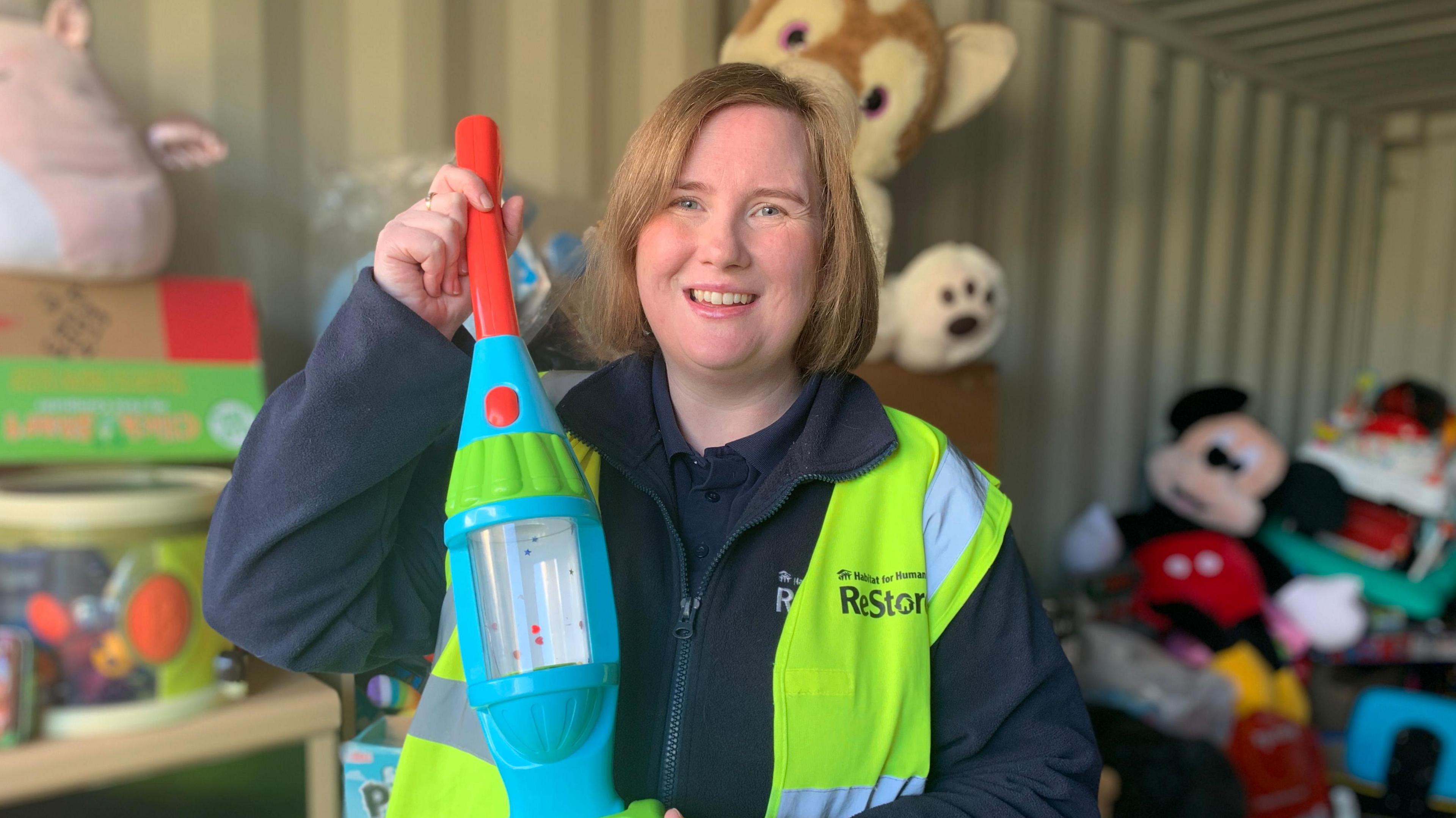 A woman with chin length light brown hair smiles toward the camera. She is wearing a navy blue fleece jacket and a yellow and silver hi visibility vest on top. She is holding up a toy plastic hoover. The background is blurred, but there are piles of donated toys out of focus. 