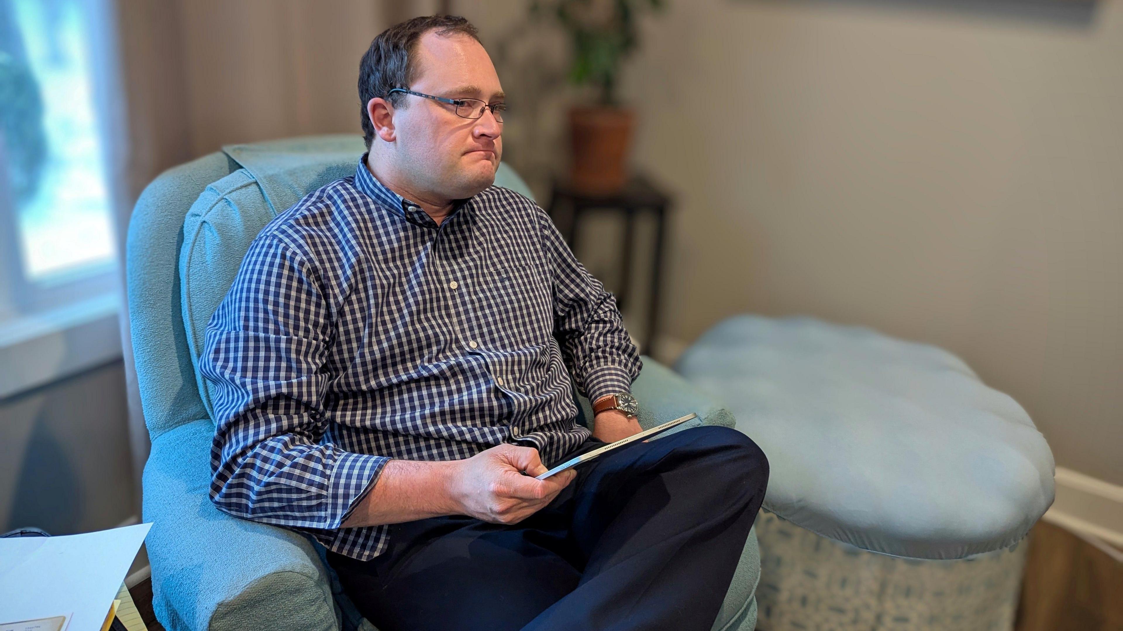 Nicholas Strahl, wearing glasses and a checked shirt, sits in an armchair holding a tablet.