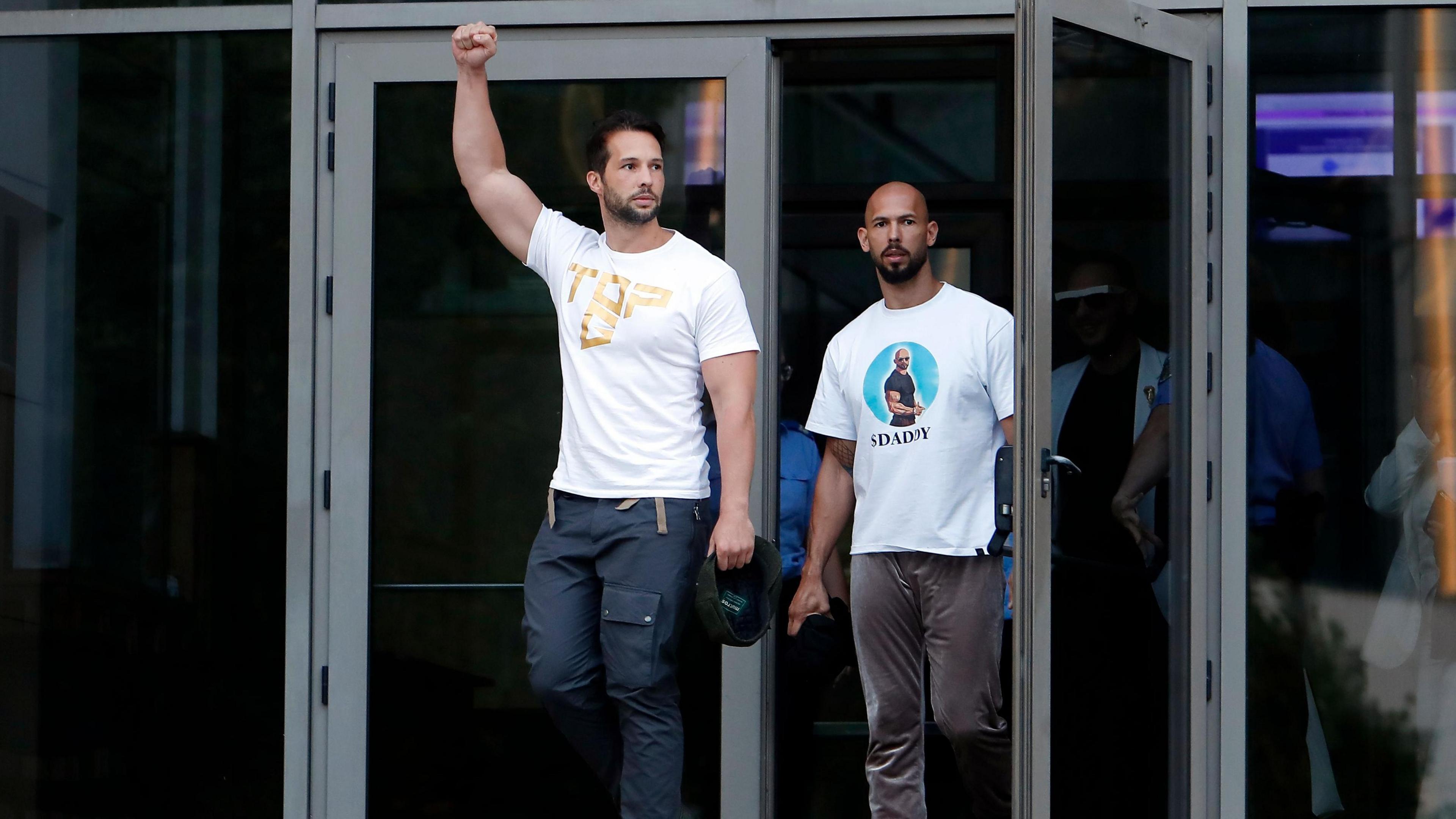 Two men in white shirts with pictures on walk out some glass doors. The man on the left, Tristan Tate, punches the air with his arm and fist.