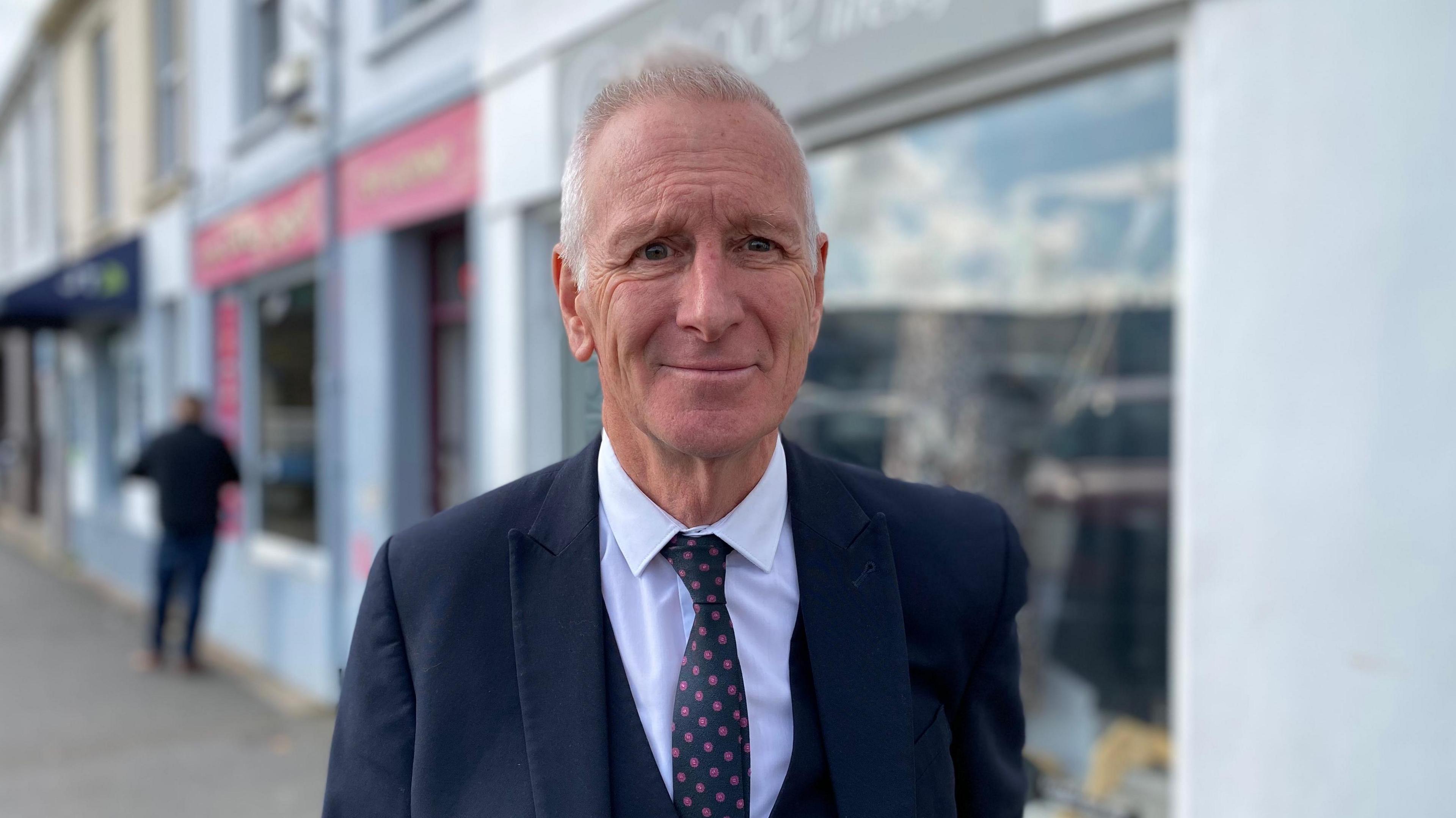 A man wearing a white shirt with a navy blue tie in a navy blue suit stands outside some shops in Guernsey.