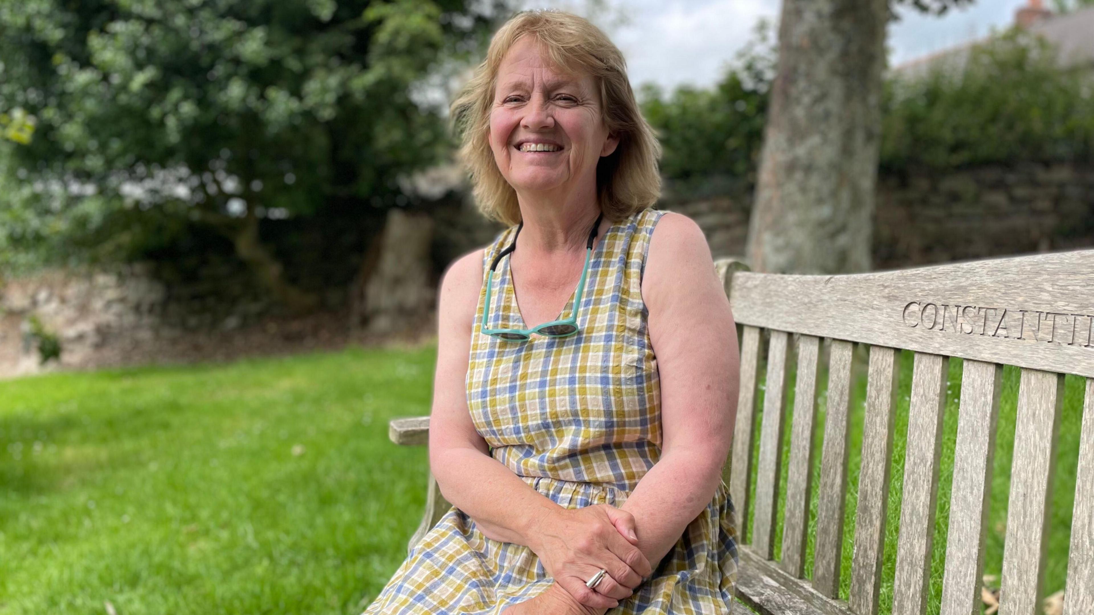 A woman in a green and blue check dress sits on a bench and smiles