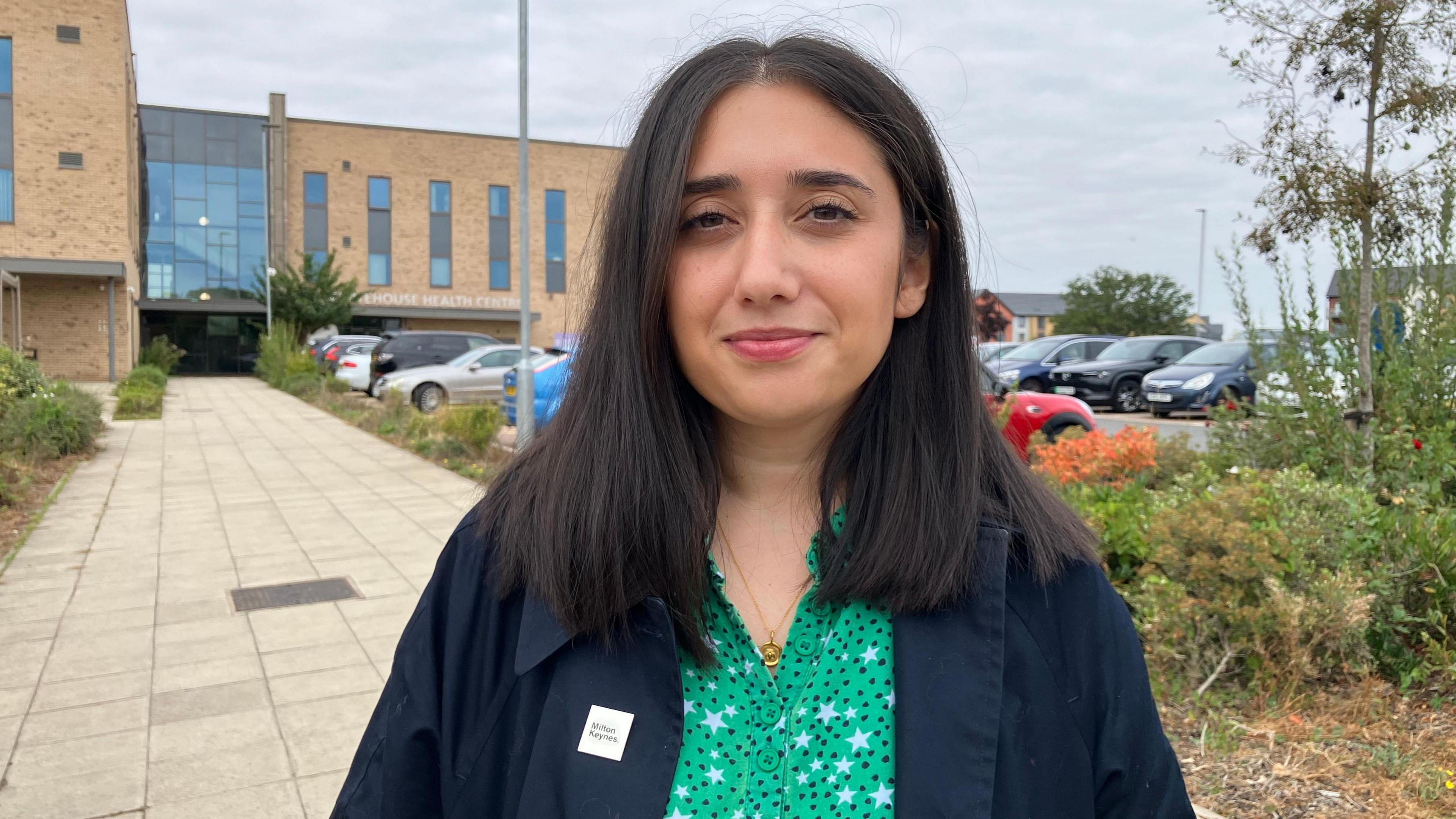 A head and shoulders shot of Shanika Mahendran who is looking at the camera and smiling. She is wearing a green top with spots on and a dark blue coat. Behind her is Whitehouse Medical Centre, which is a brown brick building with a large window above the entrance. A car park, a large paving slab pathway and some bushes can also be seen.