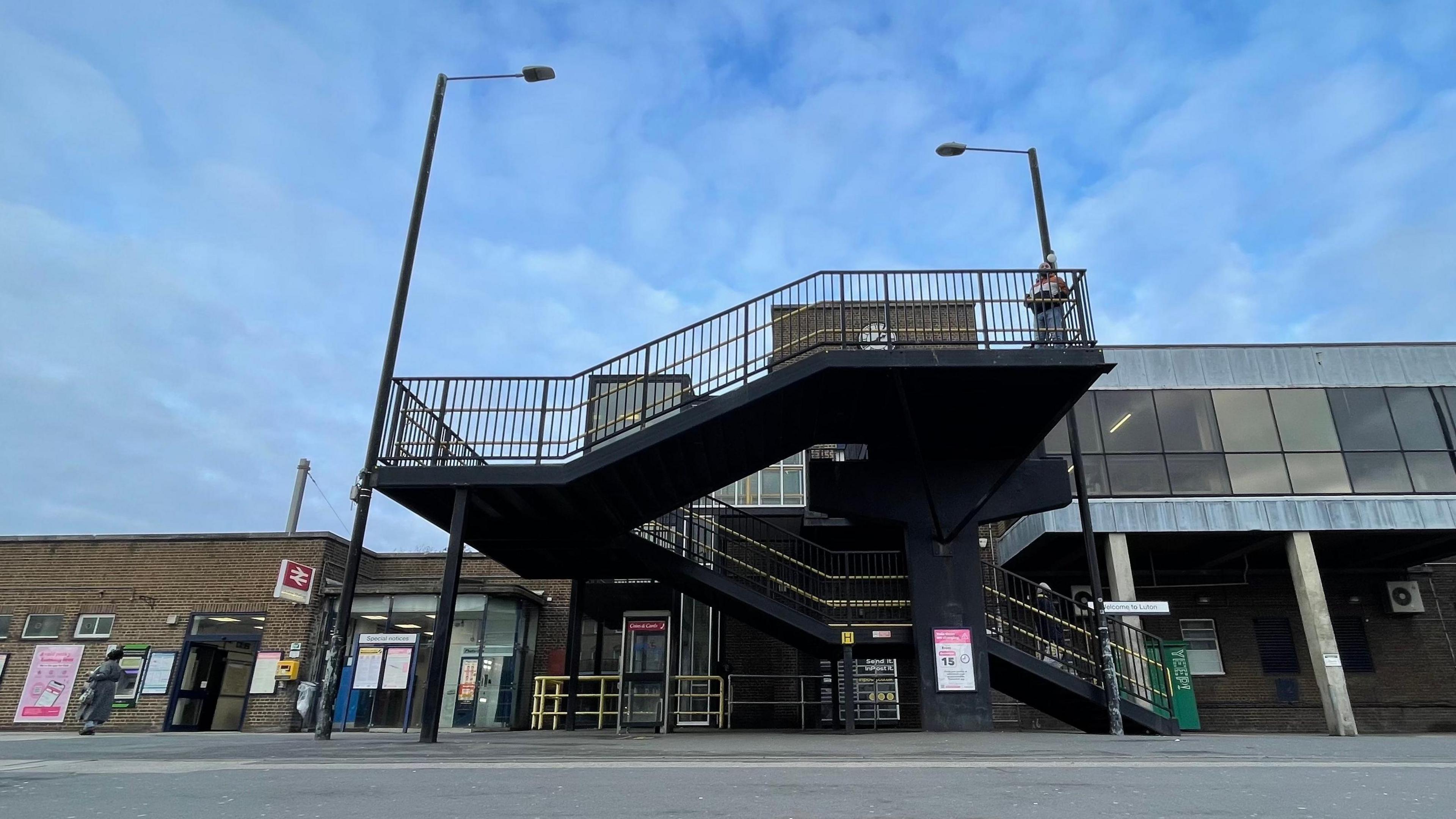 The exterior of Luton Station, as seen from the town centre side of the tracks. Two lampposts stand either side of a set of stairs that dog-leg from the street level to an un-seen ticket office. A red and white railway sign can be seen on the left of the image. There is  tarmac in the foreground and a blue sky with thin clouds hangs above the station.