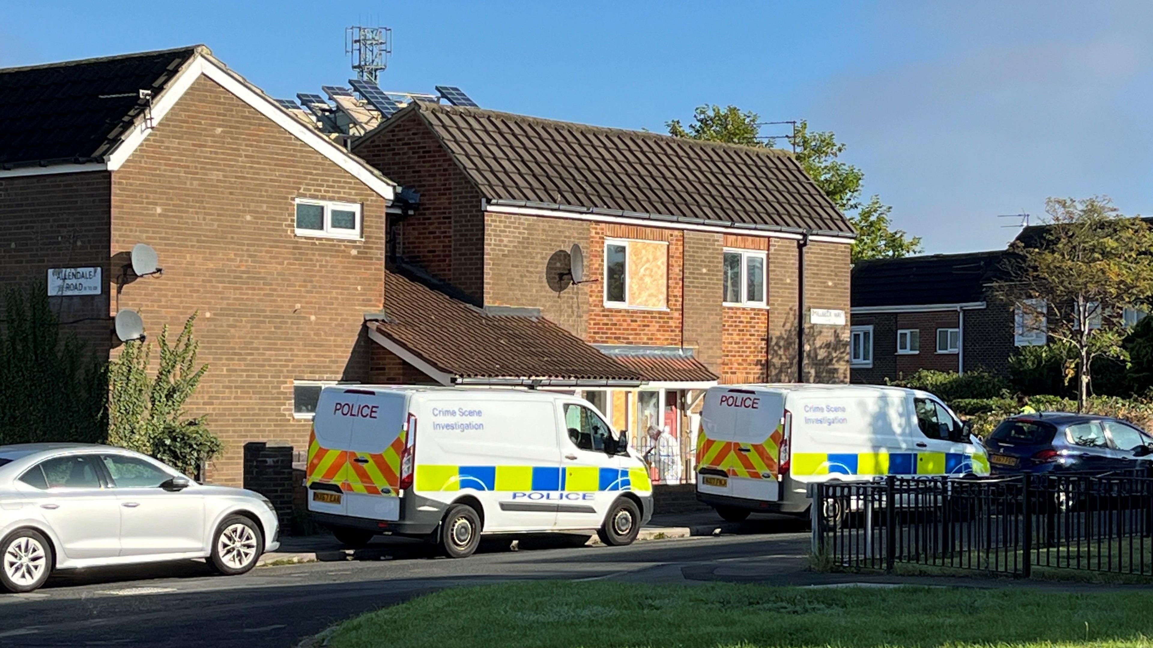Two white vans with the wording 'police' on the back and the wording 'Crime Scene Investigation' written on the side, are parked on a residential street. A person in a white suit and mask can be seen entering a house. 
