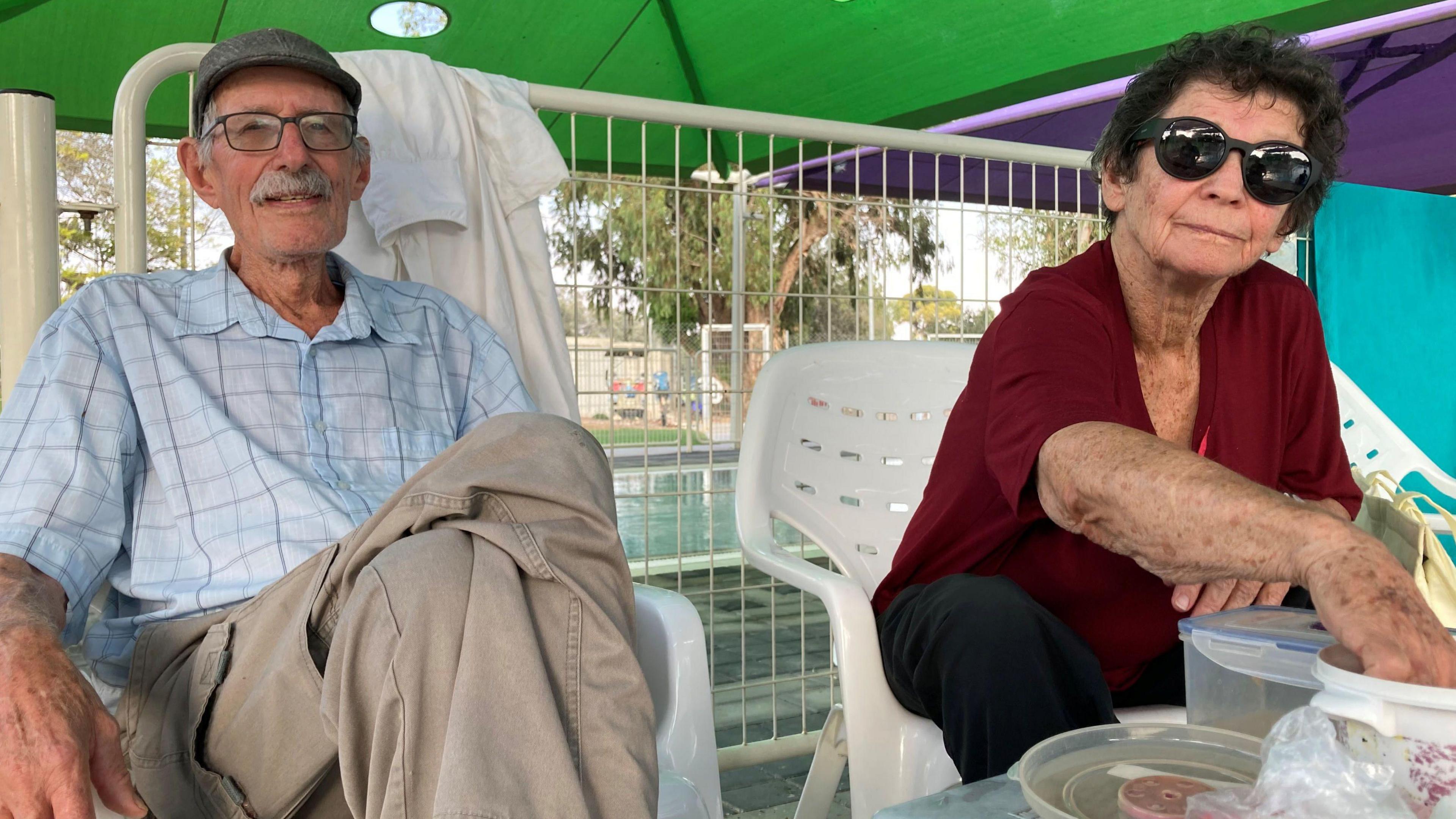 An older man and his wife sit on plastic garden furniture. 