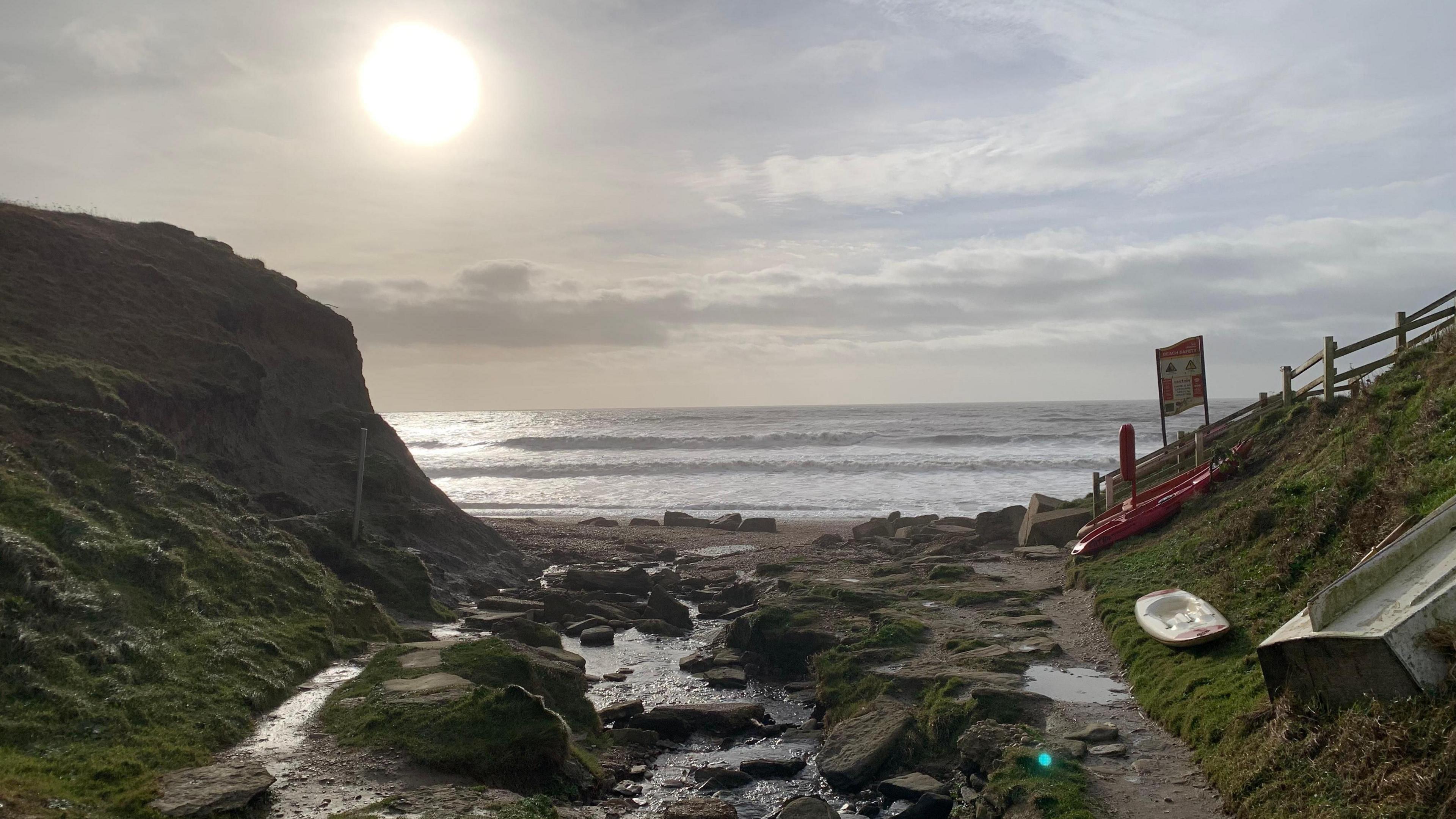 There is a wide path down to the beach with large stones and boulders lying on the ground. The path opens up on to a sandy beach where the waves are lapping gently on to the shore. A hazy sun is low in the sky.