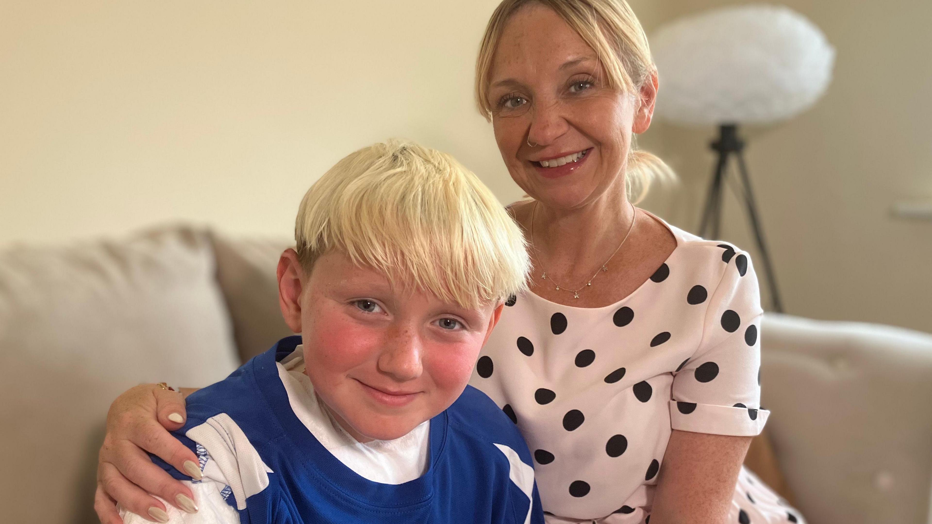 Hector and Natalie sitting on a sofa at home. They both have blonde hair and are smiling. He is wearing a blue football shirt. She is wearing a polka dot top