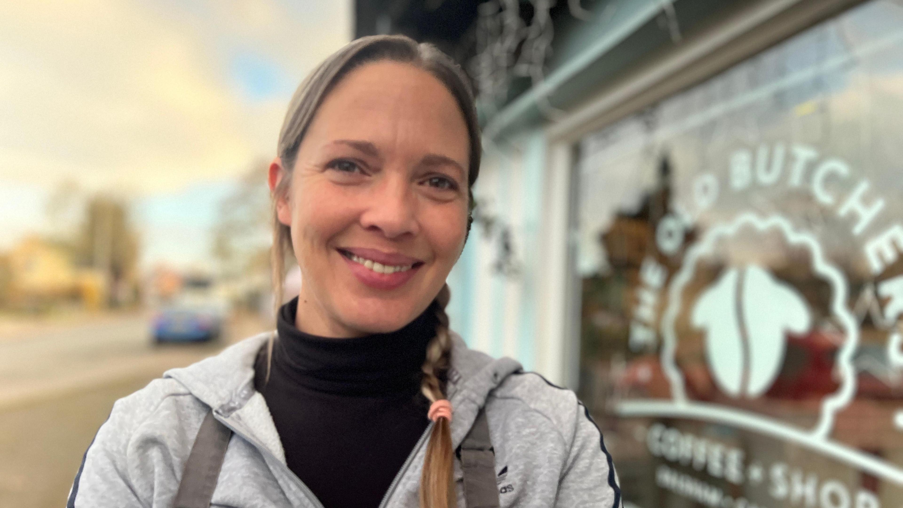 Holly Venn is wearing an apron and grey jumper, she is smiling at the camera, in the background is the glass front of her coffee shop.