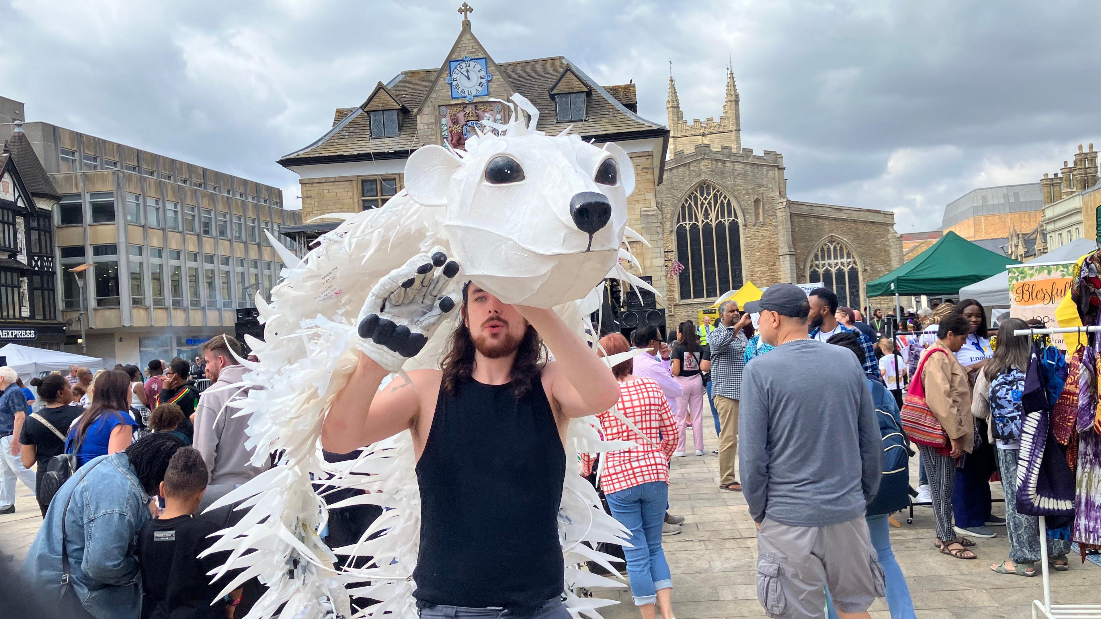 A man holding an animal head-dress, made for a carnival, in a city centre square, with people all around him. He has black singlet. There are stalls around him and buildings behind him. 