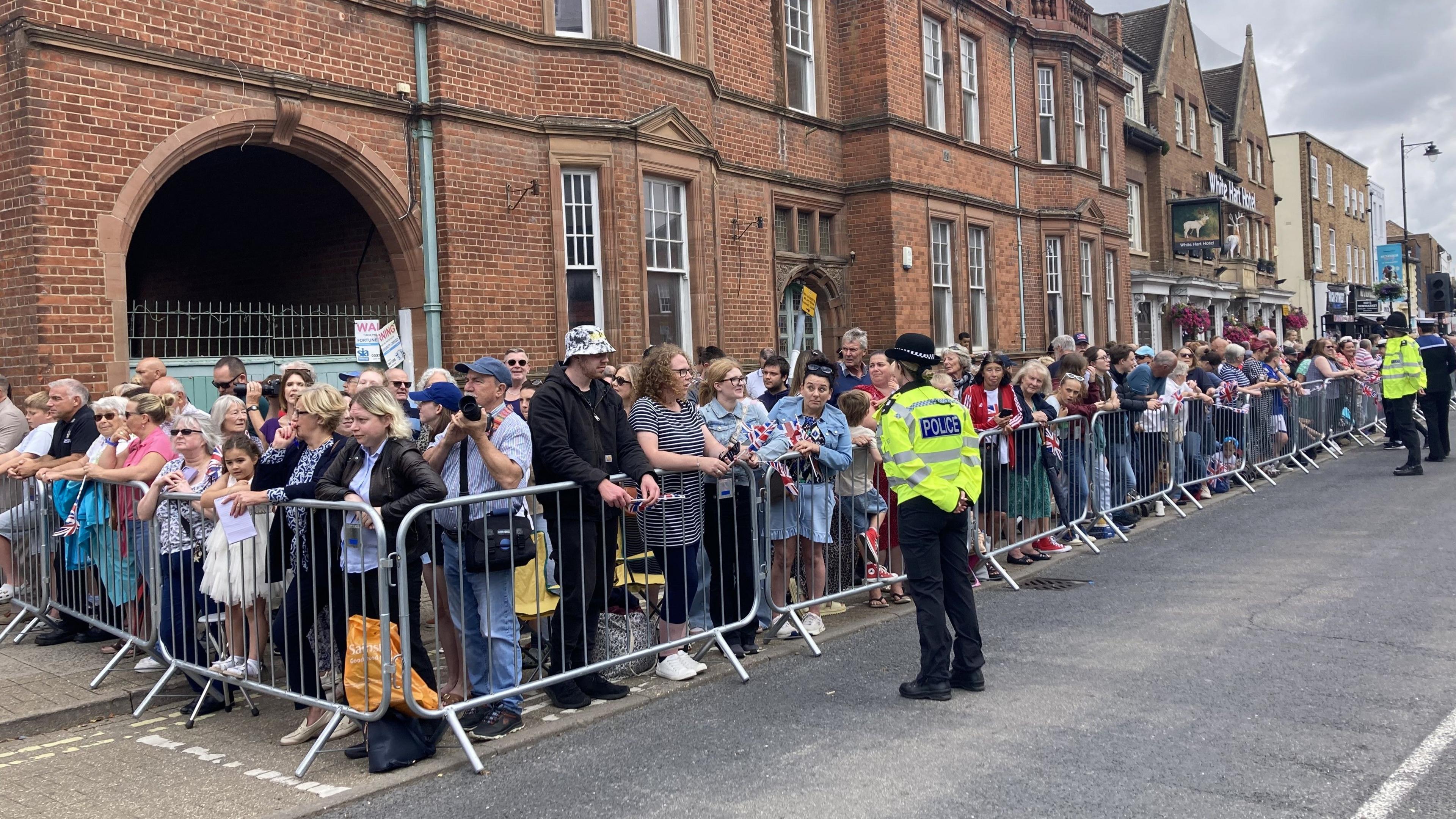 Crowds of people stand behind a barrier ready to welcome the King and Queen. Uniformed officers stand on the other side of the barrier watching the crowds.