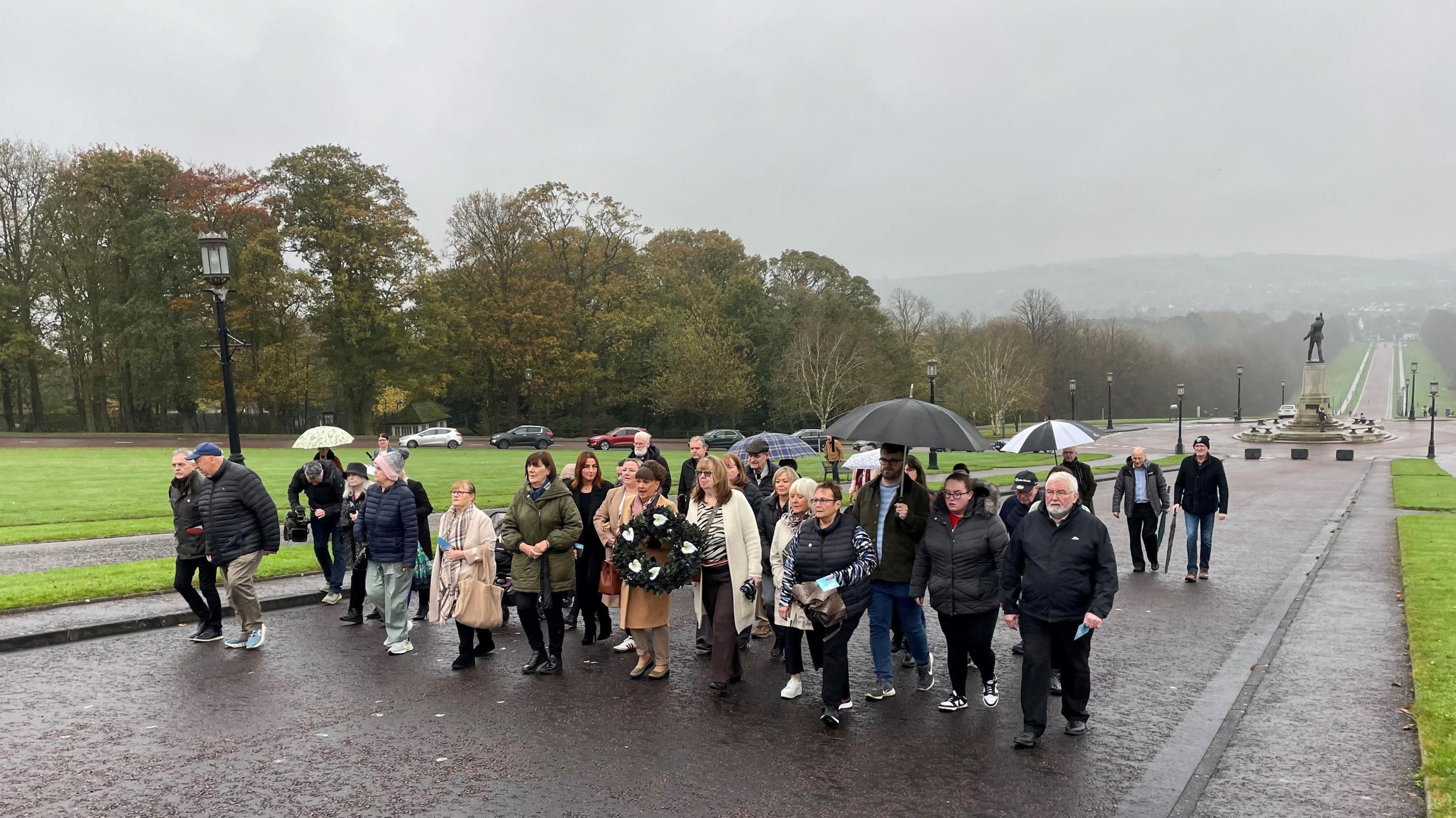 A group of people walking on red tarmac towards Stormont buildings, its raining, some have umbrellas