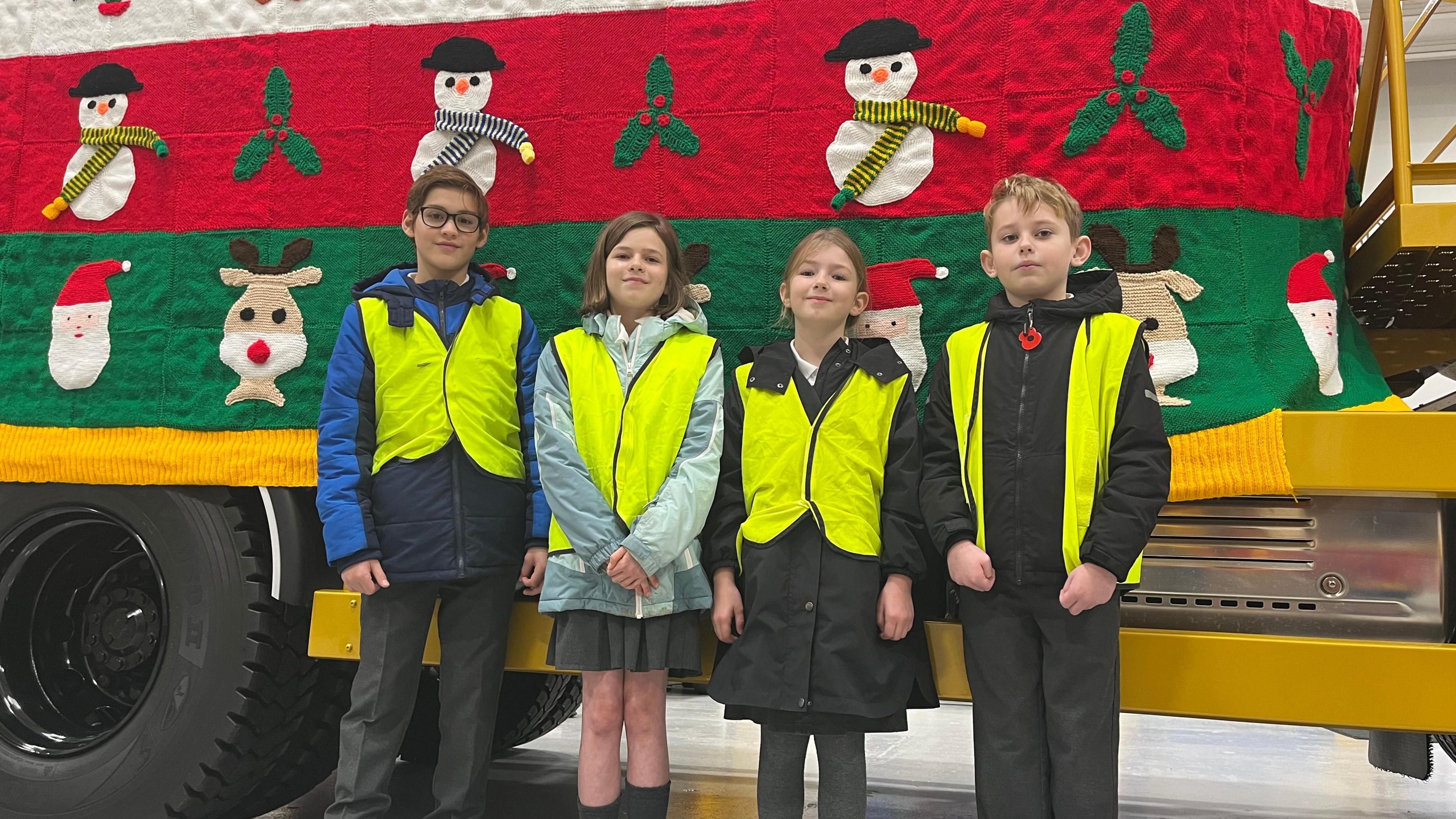 Four children in hi-vis jackets stand in front of the gritter