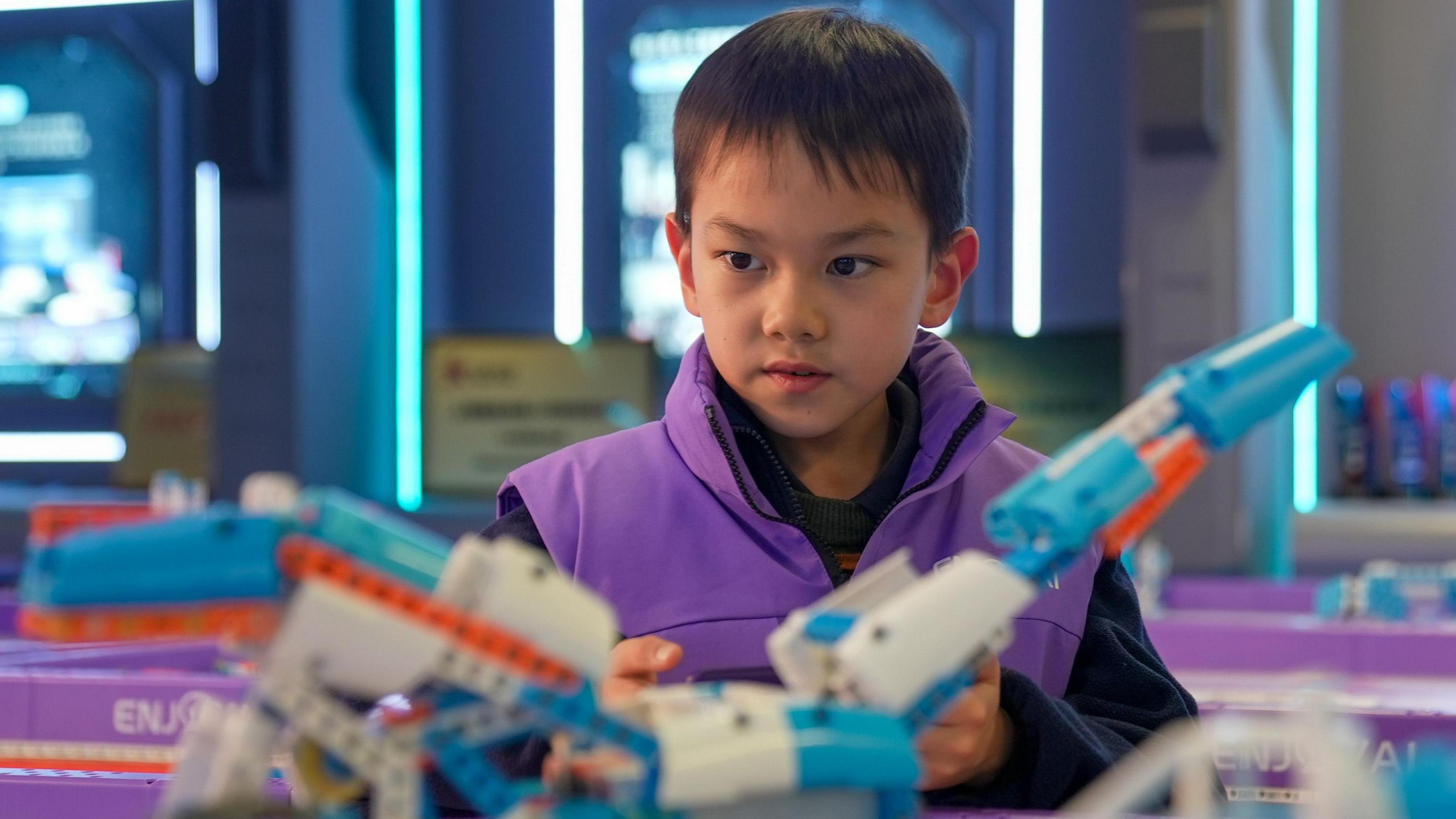 A child in a purple jacket operates a blue and orange AI-powered toy he built using code 