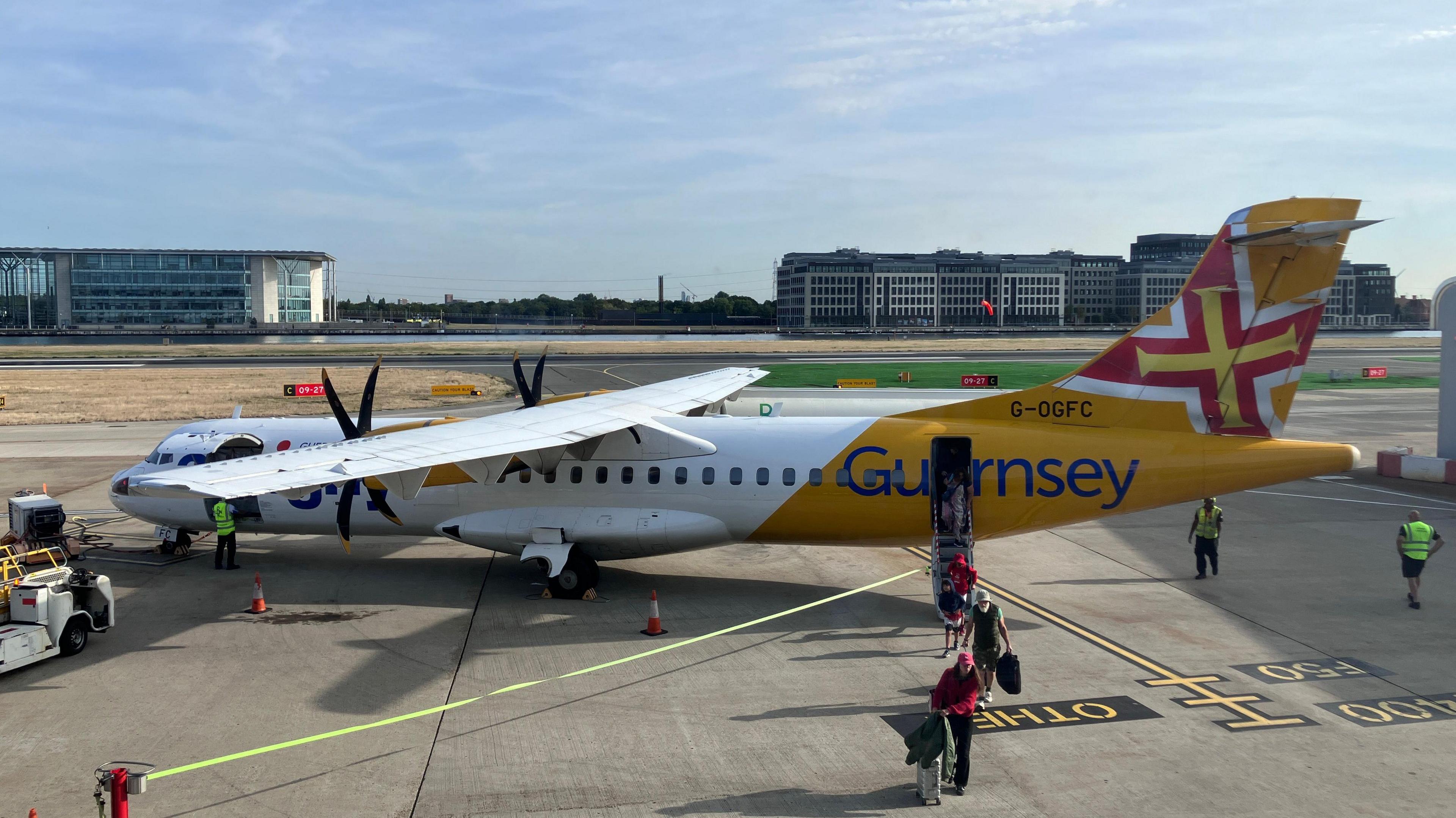 Passengers stepping off an Aurigny ATR plane at an airport, with the terminal buildings behind it. The twin-propellered plane, which can hold about 80 people, has white and yellow livery