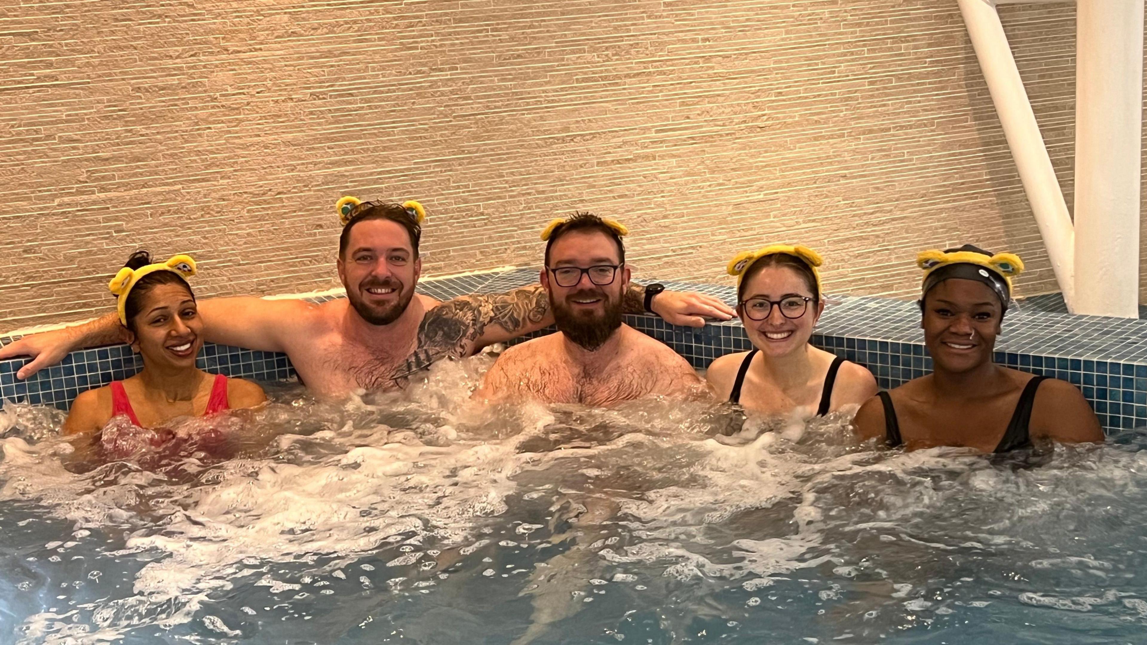 Two men and three women sitting in a line in a hot tub, all wearing yellow Pudsey ears and smiling at the camera.