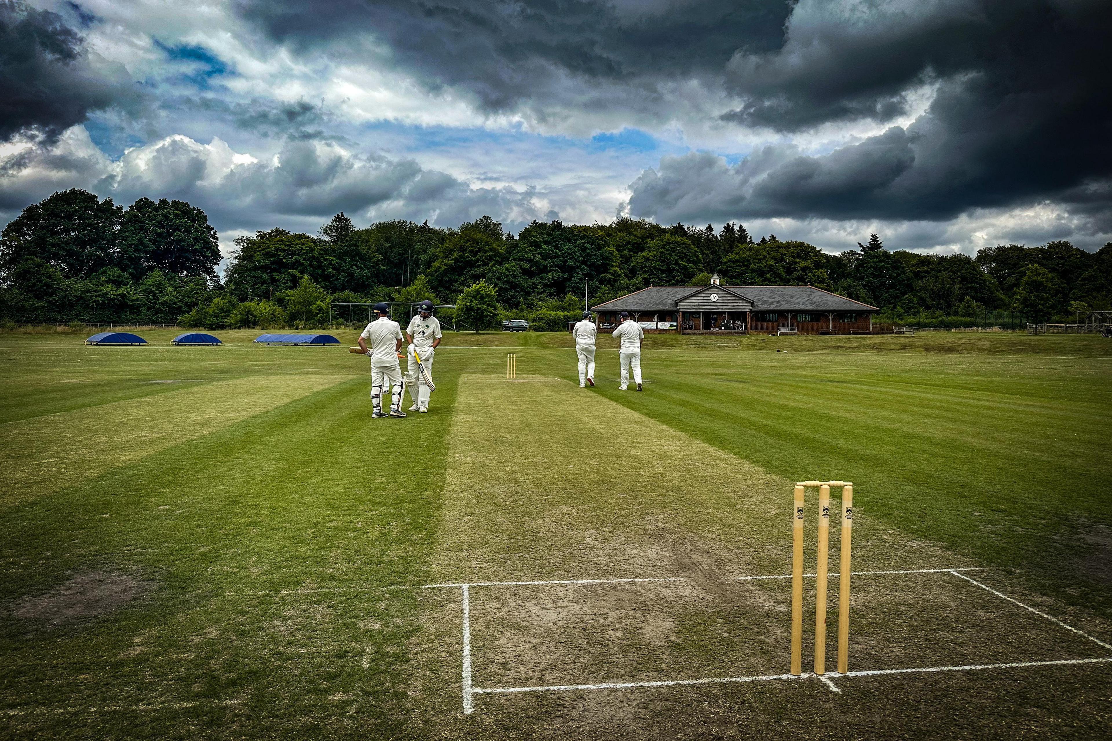 A cricket square with stumps and white lines dominate the image with two batsmen to the left of it in discussion. On the right of the square a bowler and his captain can be seen walking away also discussing tactics. Above the sky is menacing with dark clouds and just a glimpse of blue skies. A cricket pavilion and blue covers in front of a line of trees can be seen in the background.