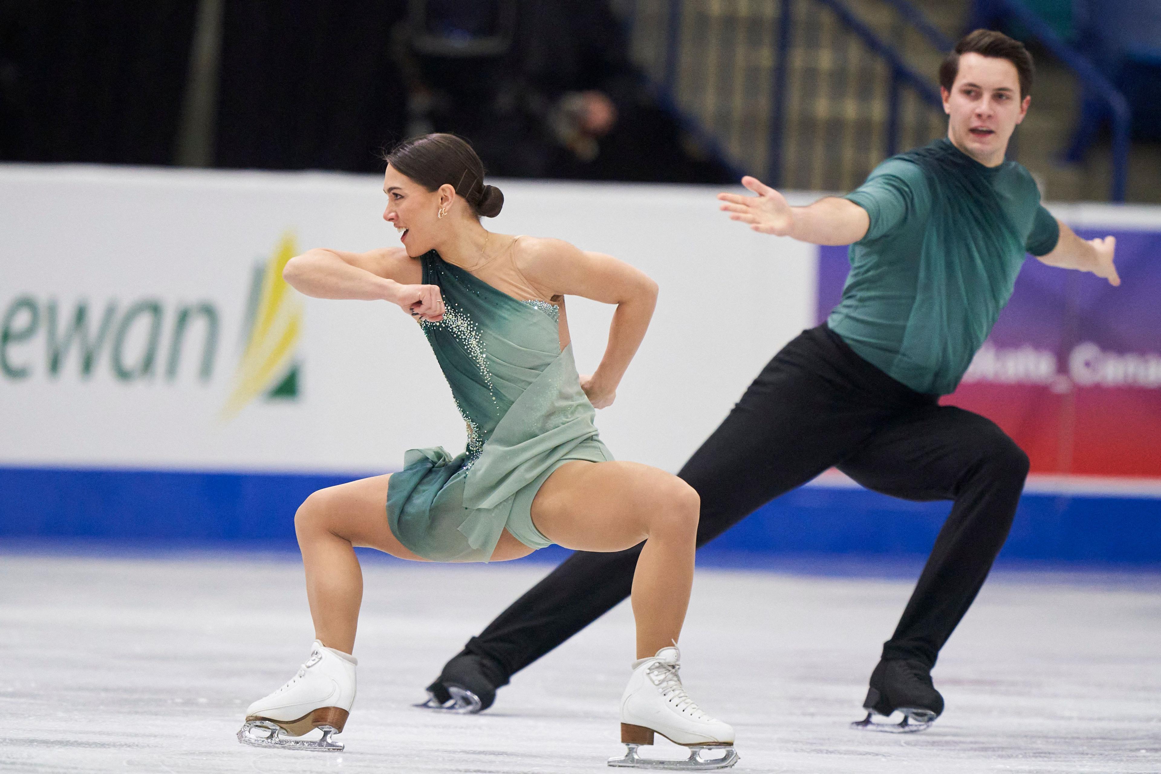 Anastasia Vaipan-Law and Luke Digby skate on the ice. Anastasia wears a dark green and light green ombre dress. Luke wears black trousers and a top that matches Anastasia's dress. 
