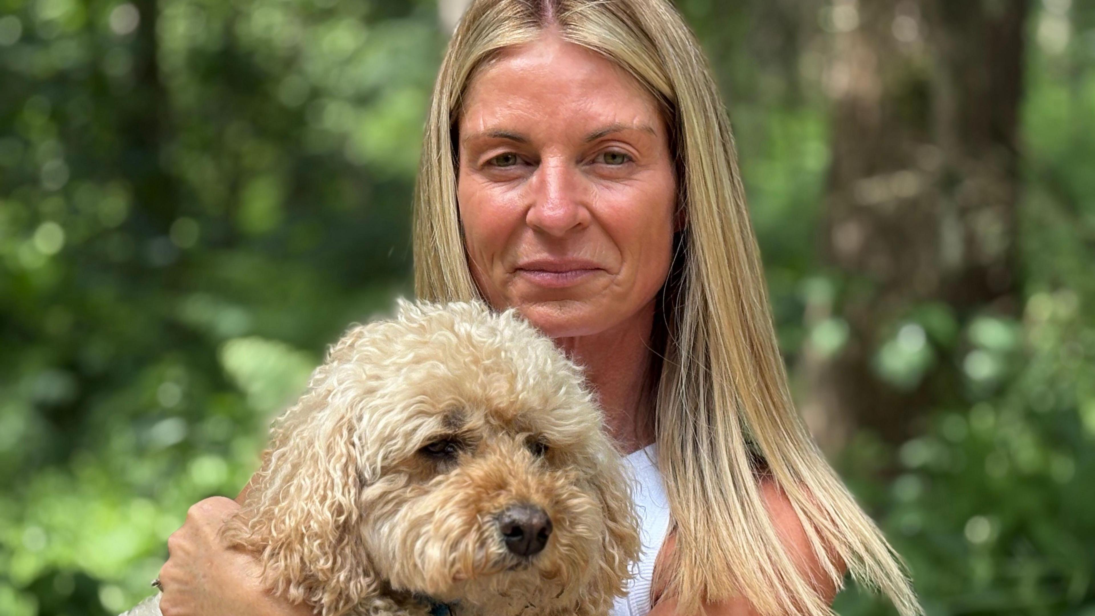 A blonde woman holds up her blonde Cavapoo dog.